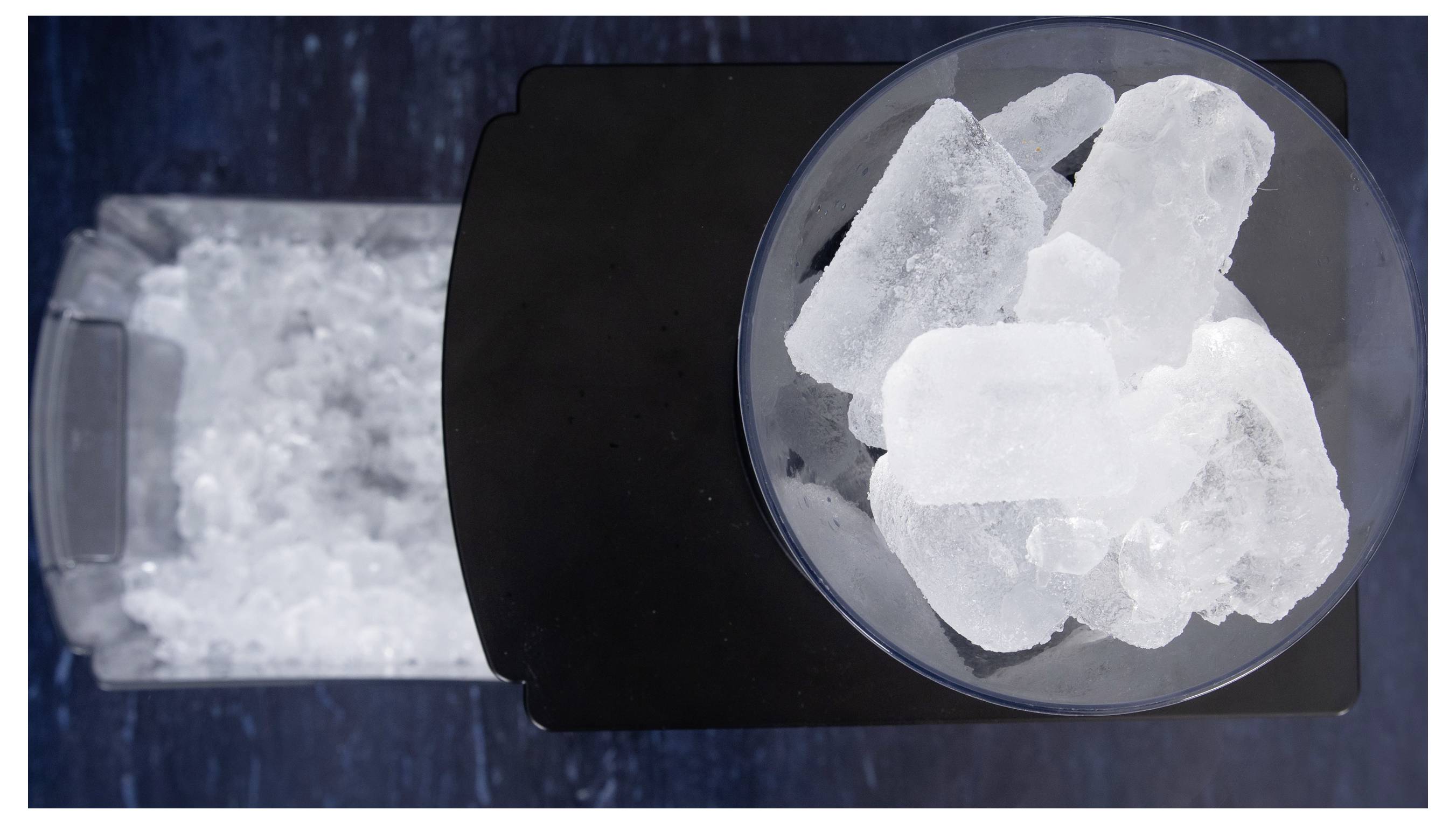 Draufsicht eines schwarzen Eismachers mit einem durchsichtigen Eimer, gefüllt mit kleinem zerkleinertem Eis, neben einer Schüssel mit großen Eiswürfeln.