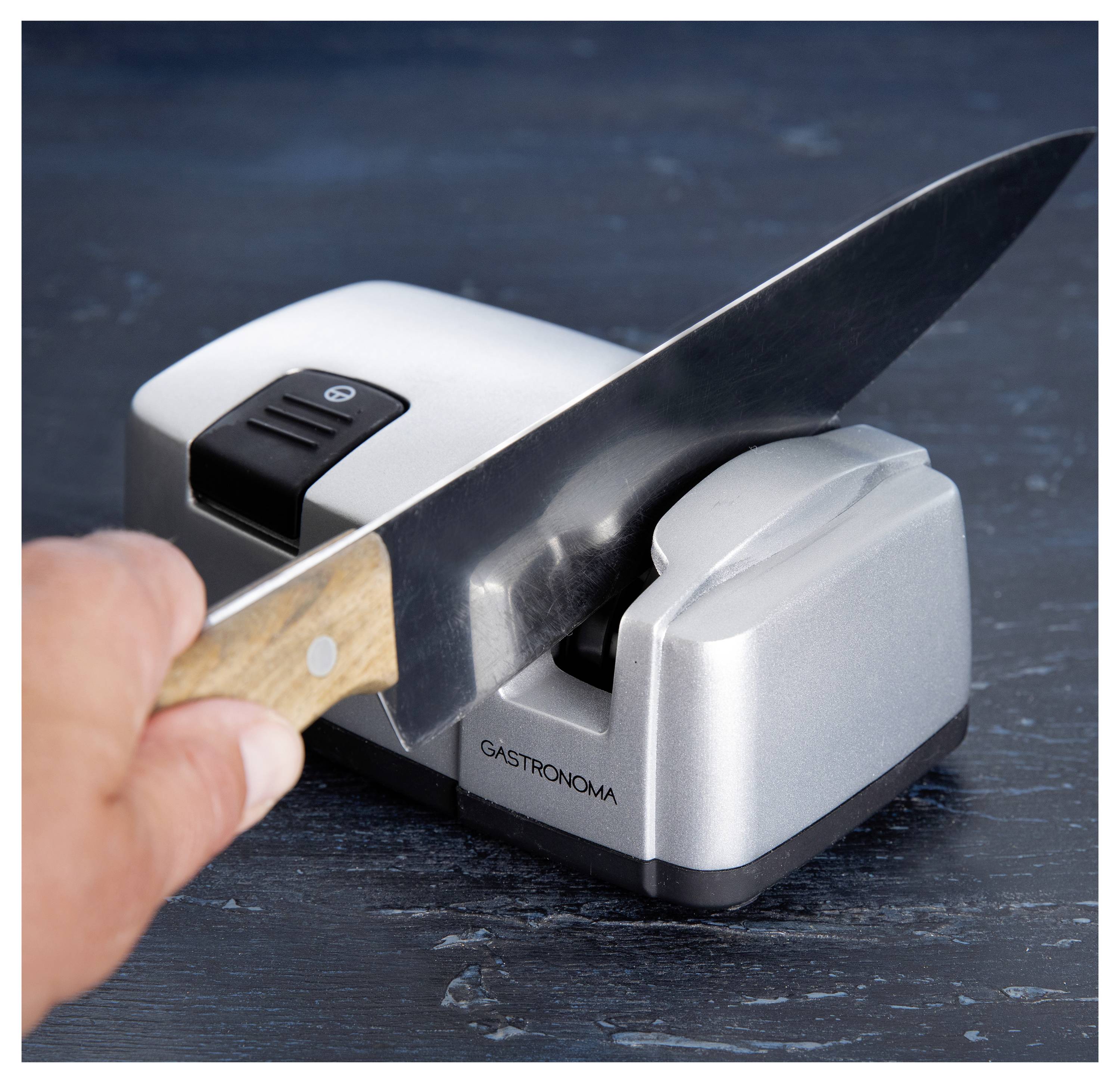 Eine Hand schärft ein Messer mit einem silbernen elektrischen Messerschärfer auf einer dunklen Oberfläche.