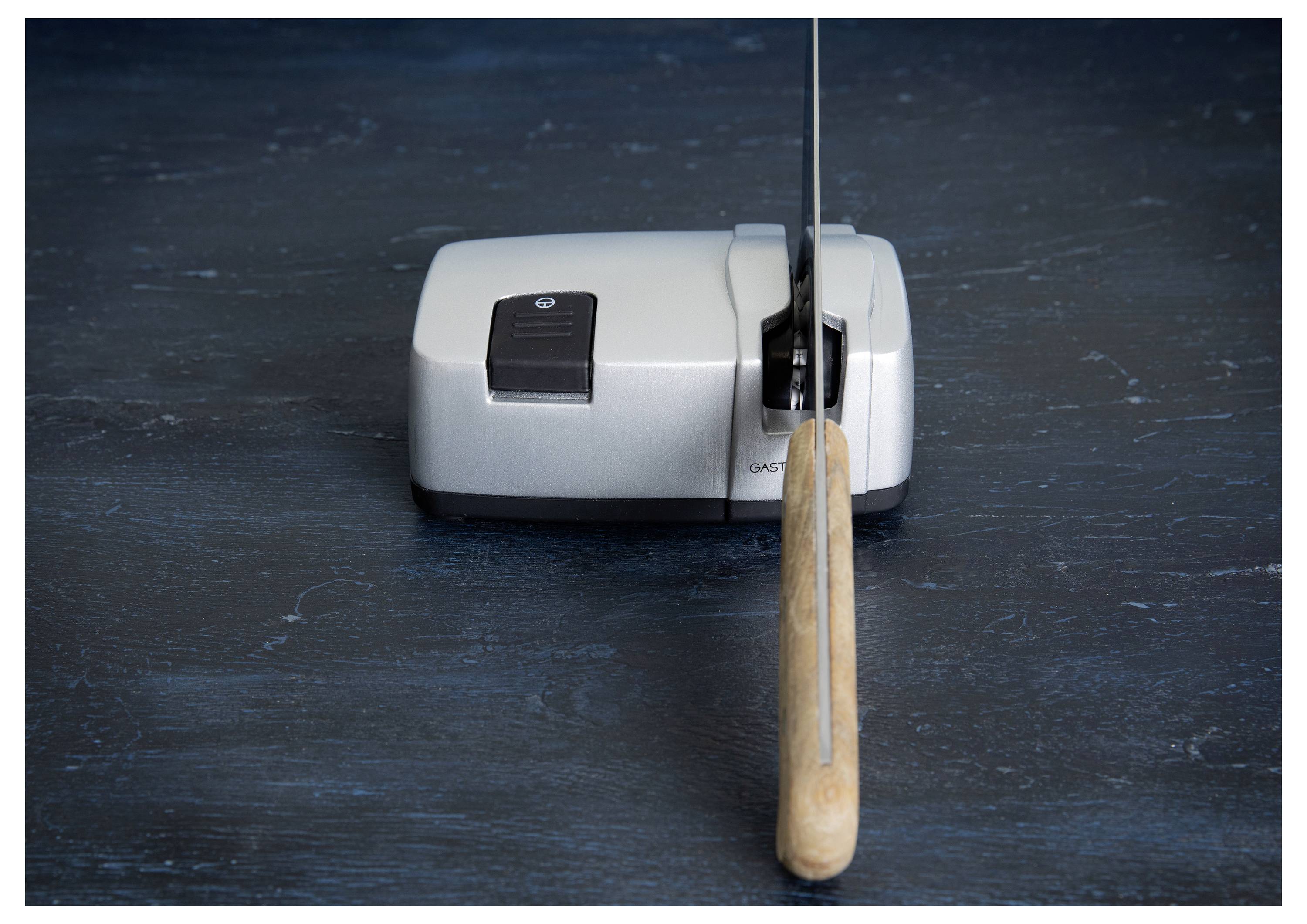Ein elektrischer Messerschärfer wird auf einer dunklen Oberfläche gezeigt, während er ein Messer mit einem Holzgriff schärft.