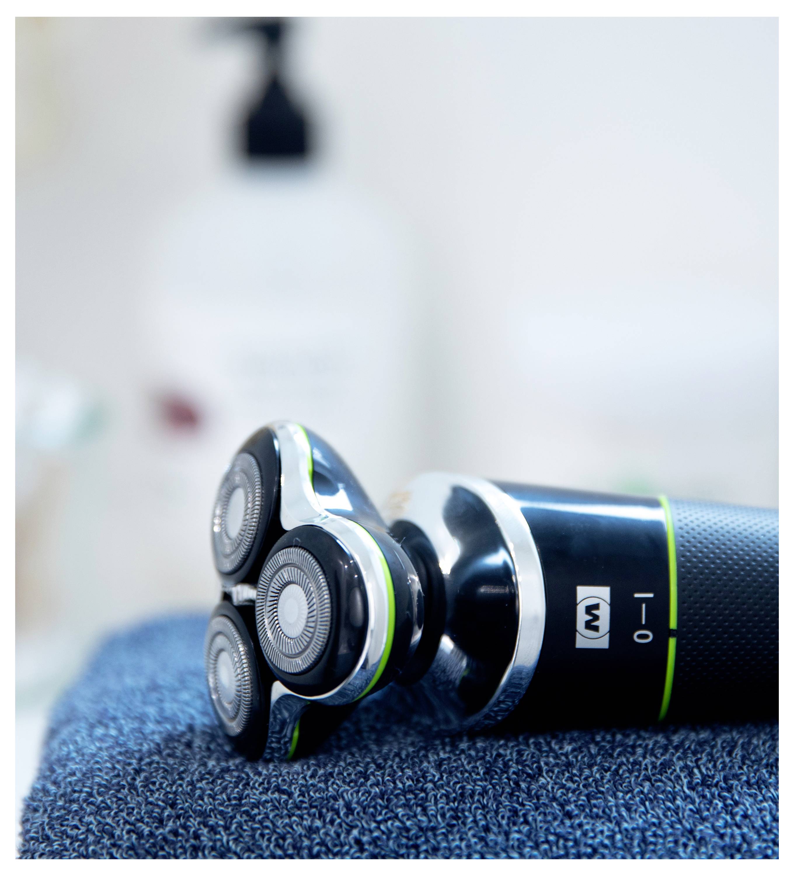 Nahaufnahme eines elektrischen Rasierers mit drei rotierenden Köpfen, der auf einem blauen Handtuch liegt. Im Hintergrund sind unscharfe Badezimmerutensilien zu sehen.