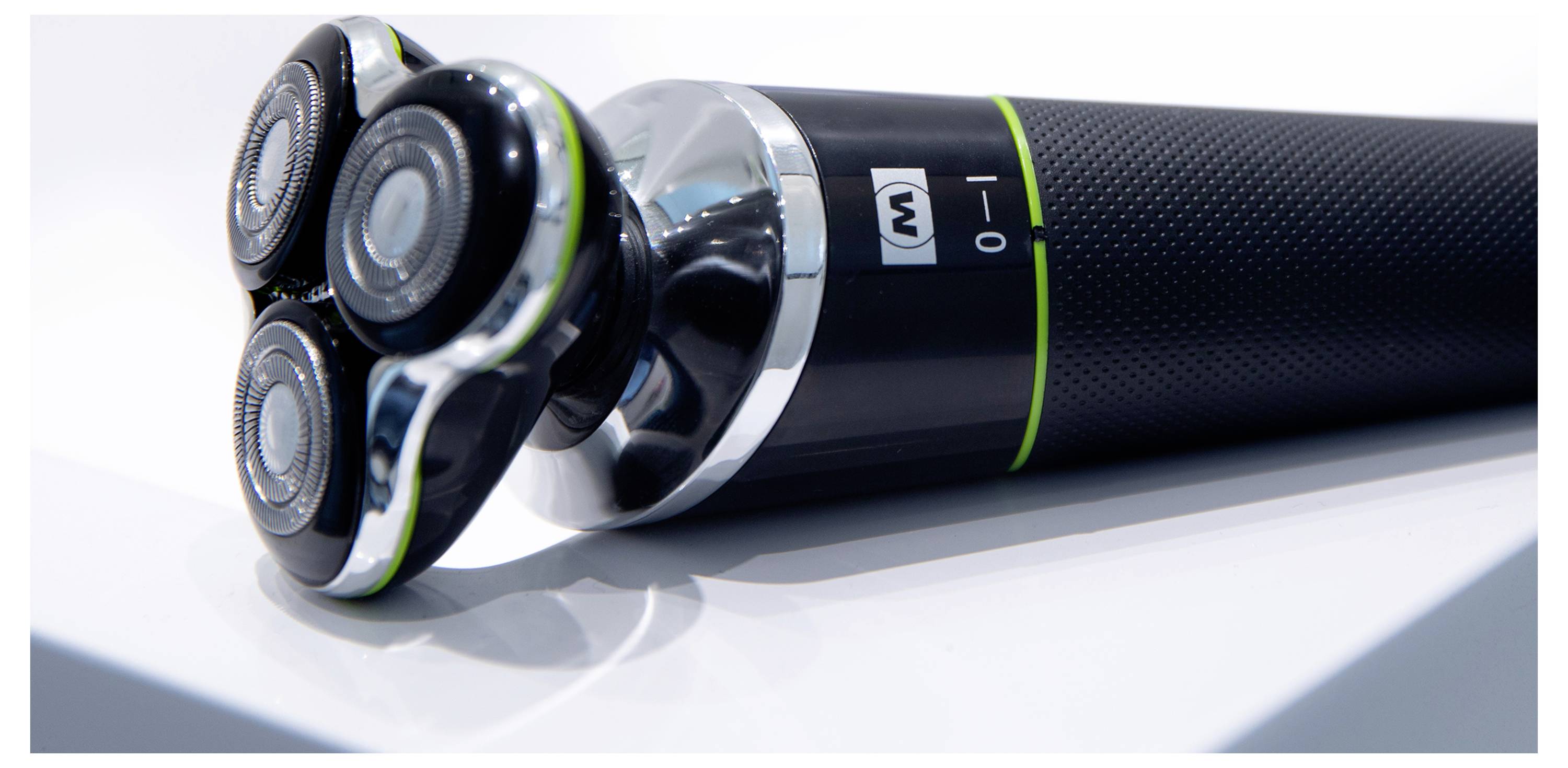 Eine Nahaufnahme eines elektrischen Rasierers mit drei rotierenden Klingen, einem Einschaltknopf und einem strukturierten Griff auf einer weißen Oberfläche.