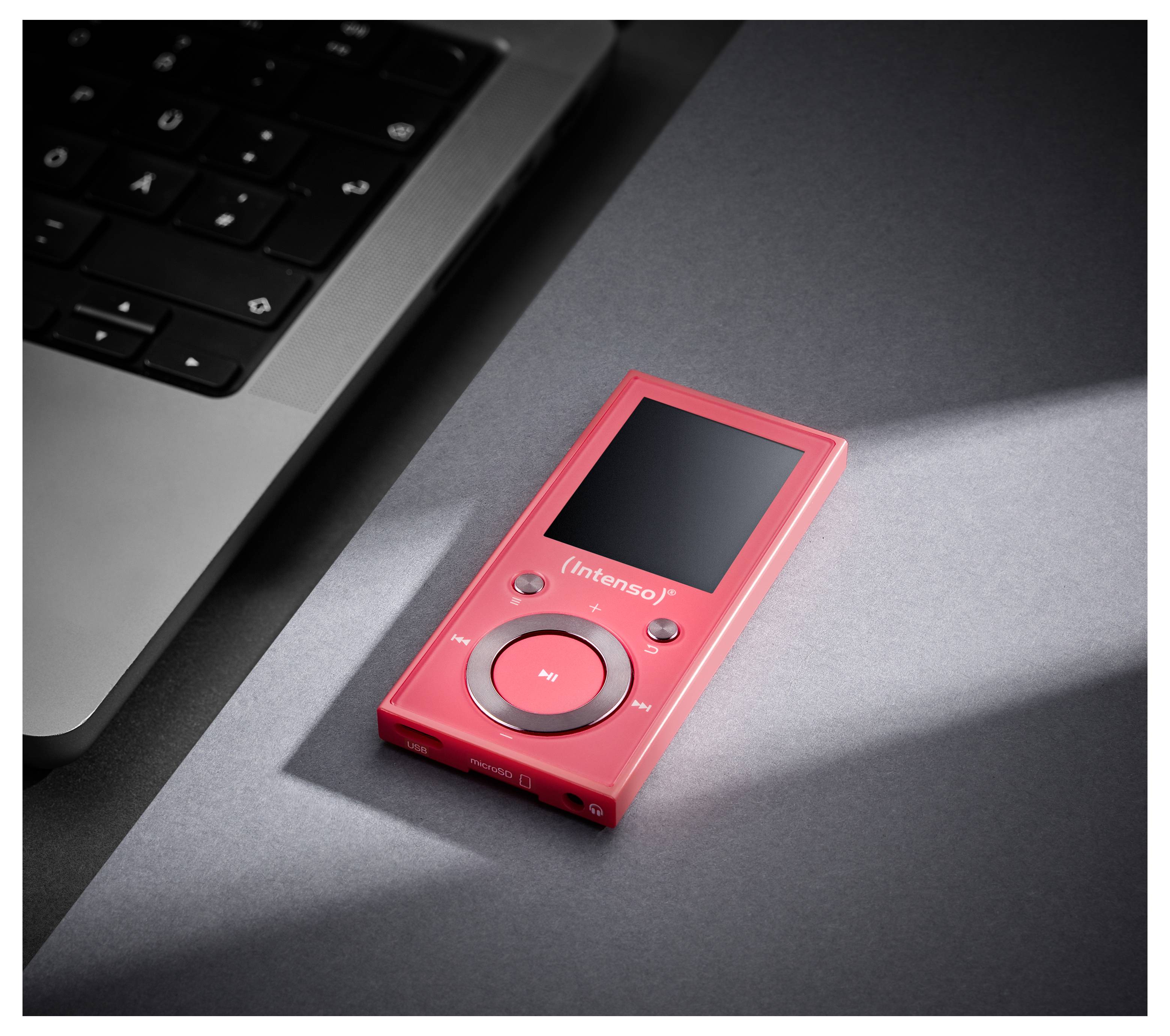 Ein roter MP3-Player der Marke 'Intenso' liegt auf einer dunklen Oberfläche neben einer Laptop-Tastatur, sanft beleuchtet.