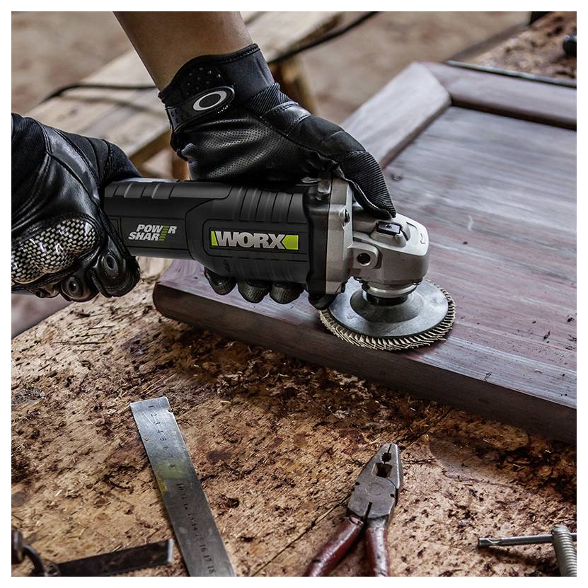 Eine Person mit Handschuhen benutzt ein Worx-Elektrowerkzeug zum Schleifen der Kante eines Holzrahmens auf einer Werkbank, die mit Werkzeugen und Holzspänen bedeckt ist.