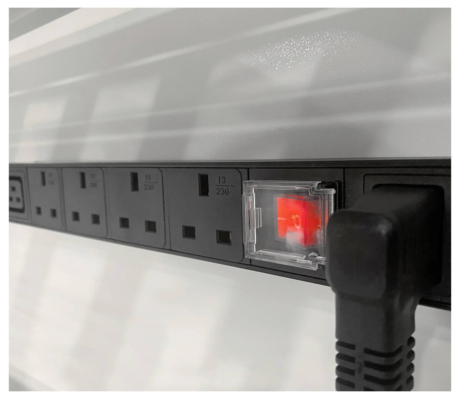 Eine Reihe schwarzer Stromsteckdosen auf einer Steckdosenleiste mit einem roten Licht, das signalisiert, dass die Stromversorgung eingeschaltet ist; ein Gerätekabel ist eingesteckt.