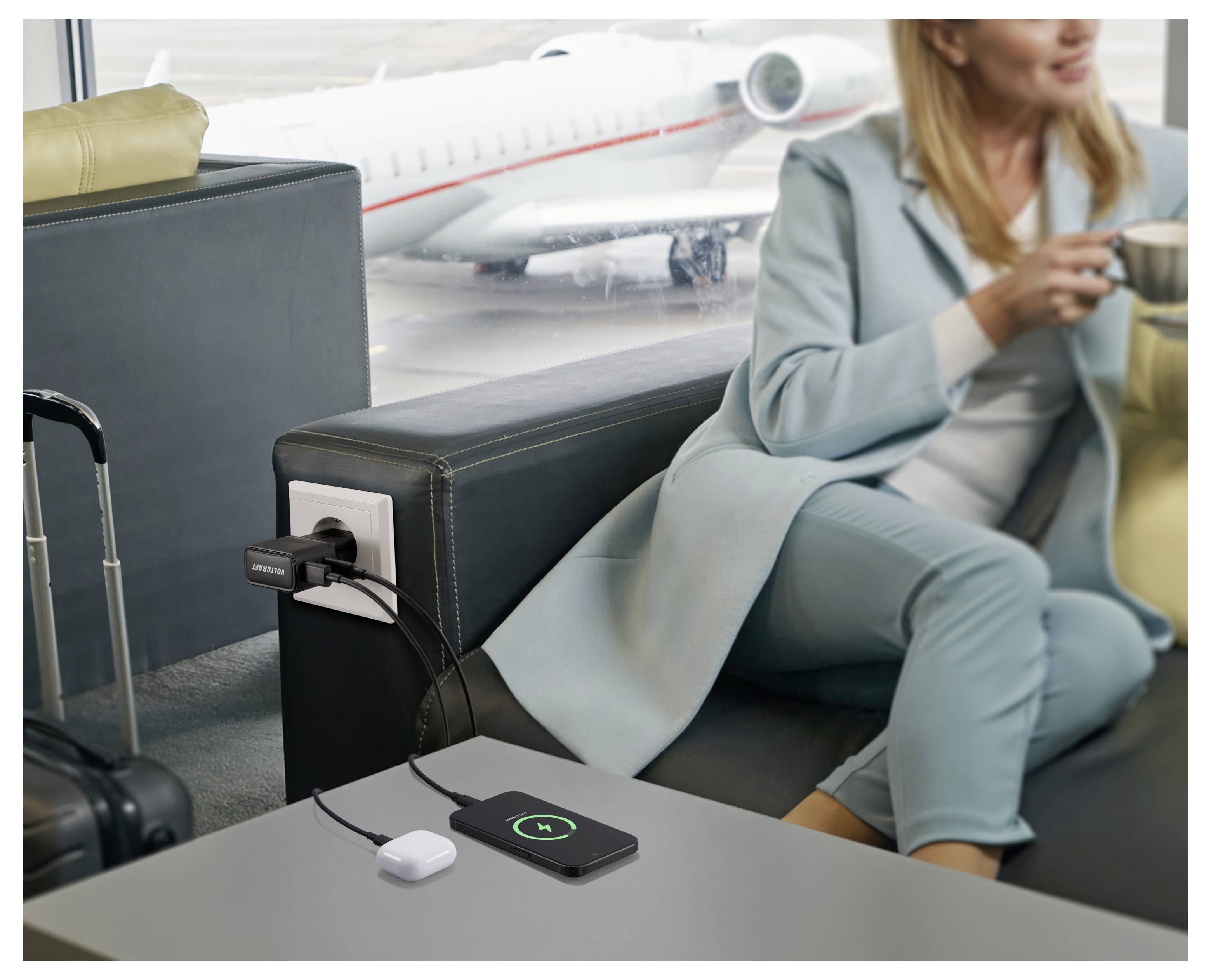 Eine Person in einem türkisfarbenen Outfit sitzt in einer Flughafenlounge und trinkt Kaffee. Ein Telefon lädt drahtlos auf einem Tisch auf, mit einem Flugzeug draußen.