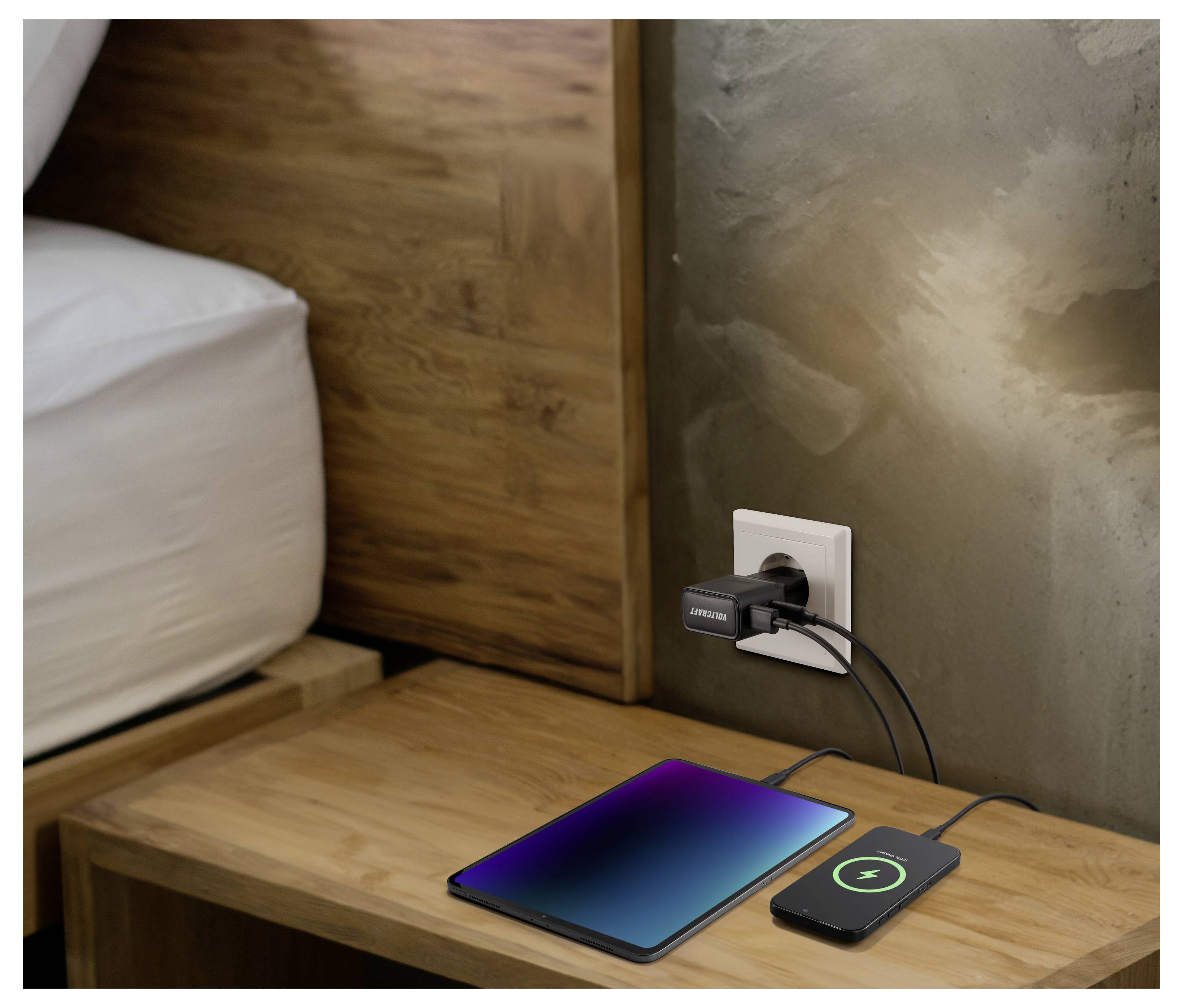 Eine Tablet und ein Smartphone werden auf einem Holznachttisch mit einem Dual-USB-Adapter aufgeladen, der in eine Steckdose neben einem Bett eingesteckt ist.