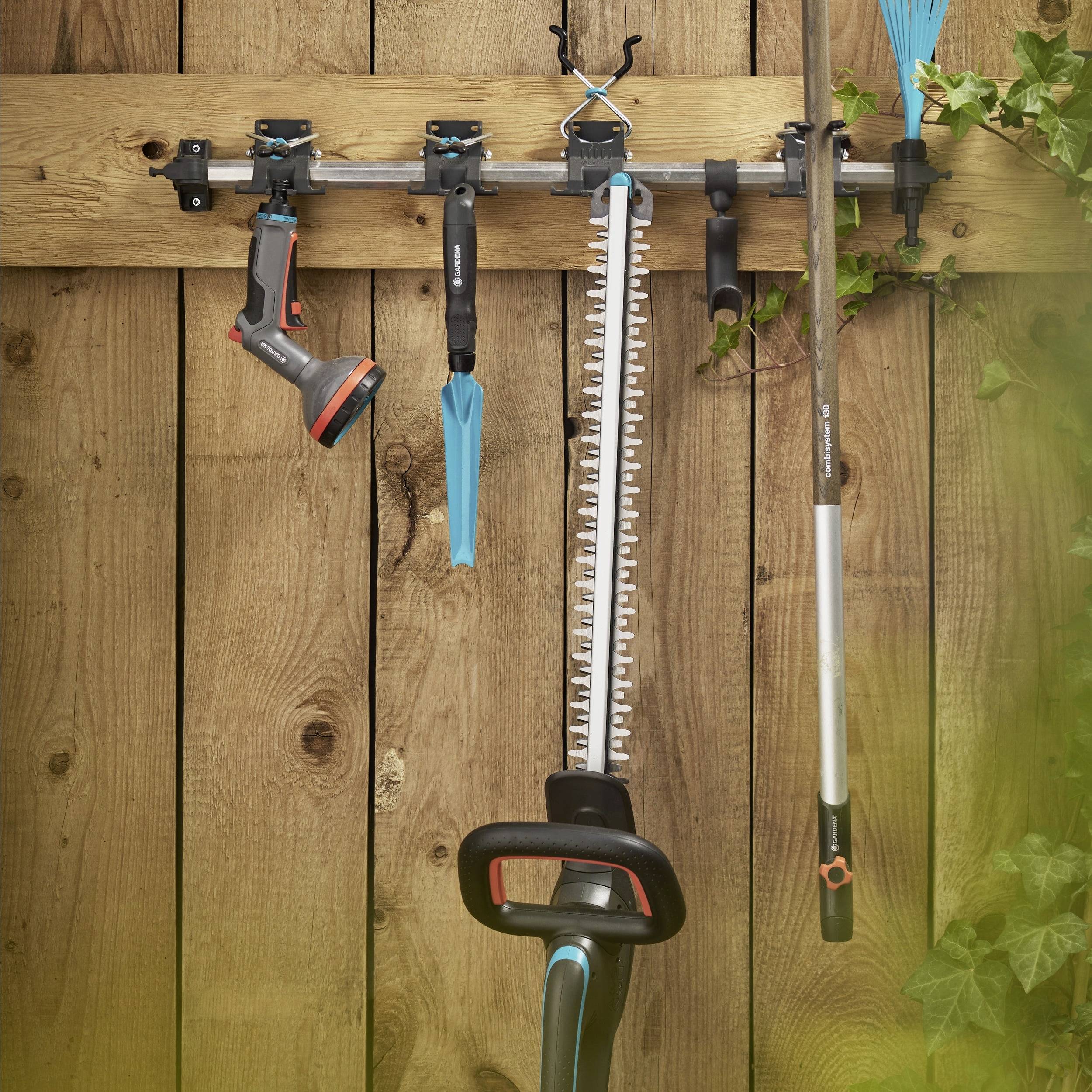Ein Holzzaun mit einem Werkzeughalter, der einen Heckenschneider, eine Gartenschlauch-Düse, einen Besen und eine Gartenschere trägt, mit Grünpflanzen im Hintergrund.
