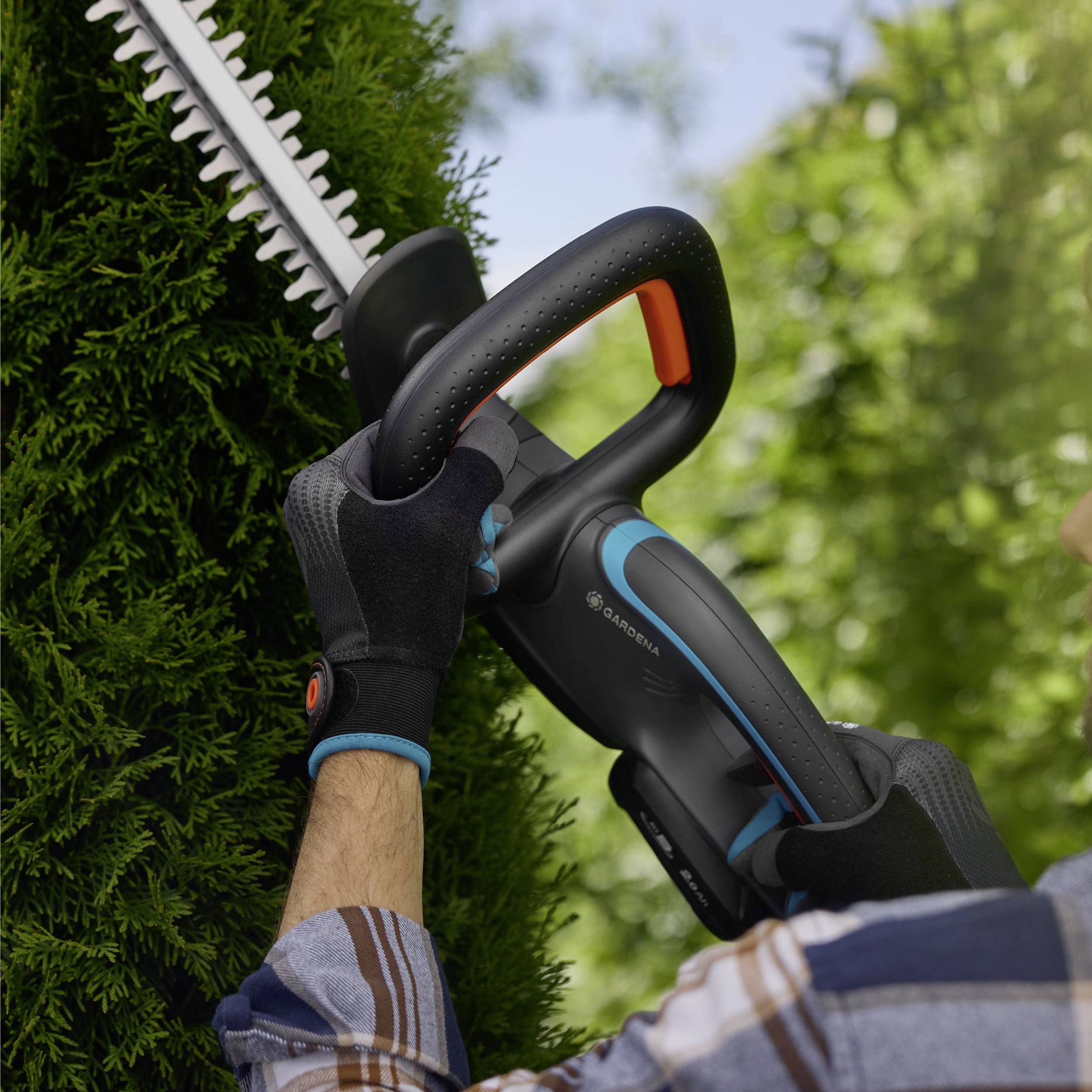 Eine Person benutzt einen kabellosen Heckenschneider, um einen Busch in einem Garten zu schneiden. Der Fokus liegt auf dem Werkzeug und dem Busch, wobei das ergonomische Design hervorgehoben wird.