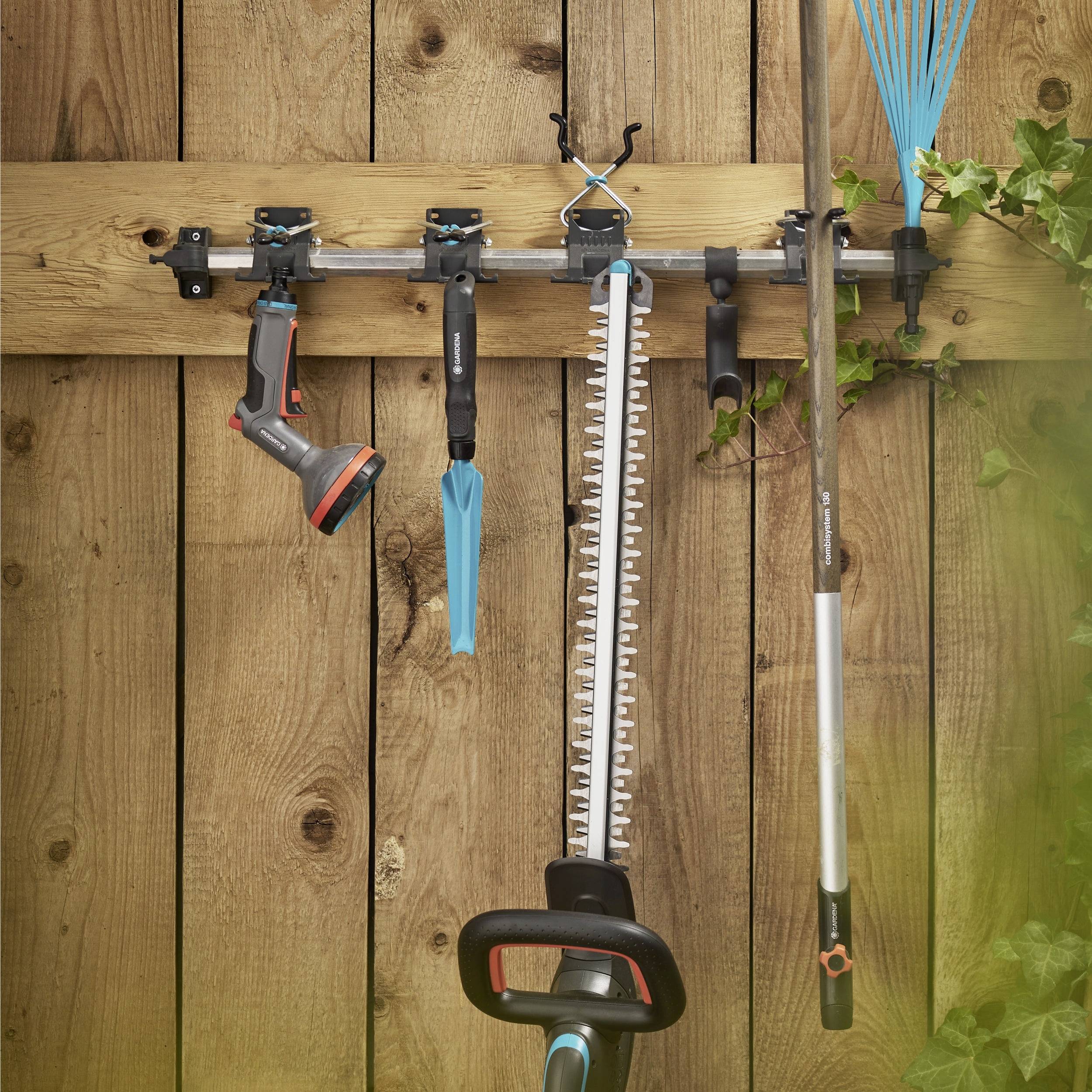 Gartengeräte, die an einem Holzregal hängen: eine Sprühbrause, Gartenscheren, eine Heckenschere, ein Rechen und Astscheren, mit Ranken in der Nähe.