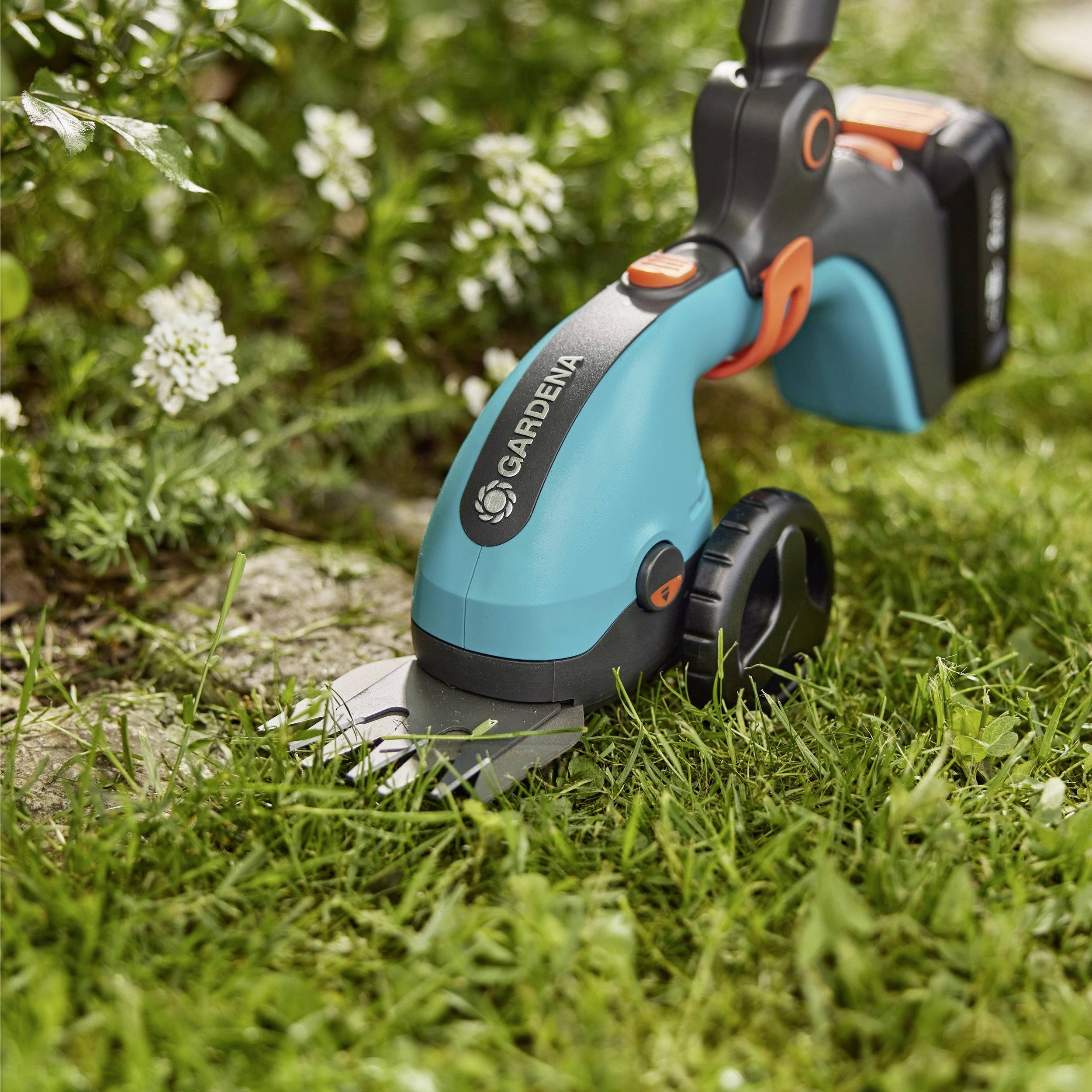 Ein blau-orangener tragbarer Rasentrimmer schneidet die Kante eines Rasens in der Nähe eines Steinweges mit weißen Blumen im Hintergrund.