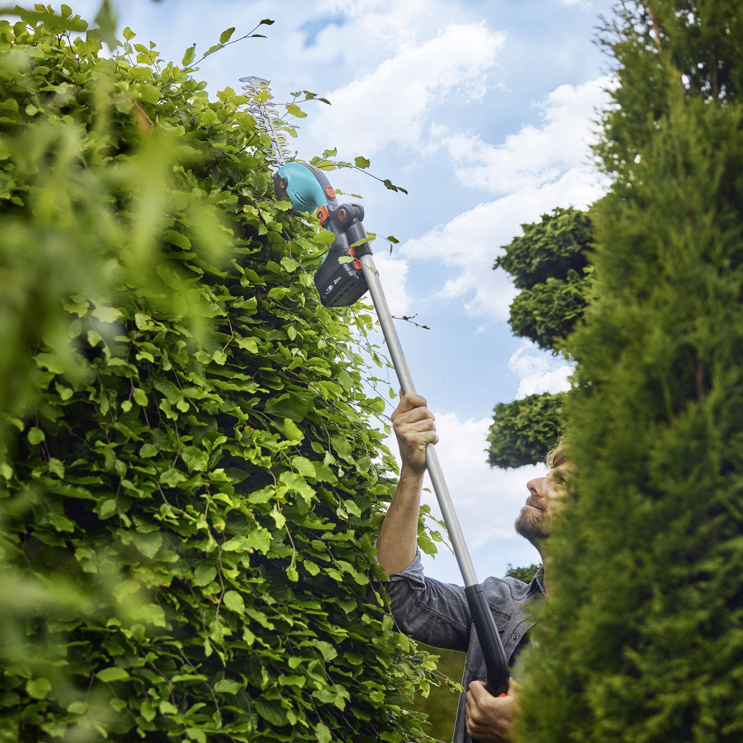 Eine Person beschneidet eine hohe Hecke mit einem elektrischen Heckenschneider an einem langen Stiel, vor einem blauen Himmel mit Wolken im Hintergrund.