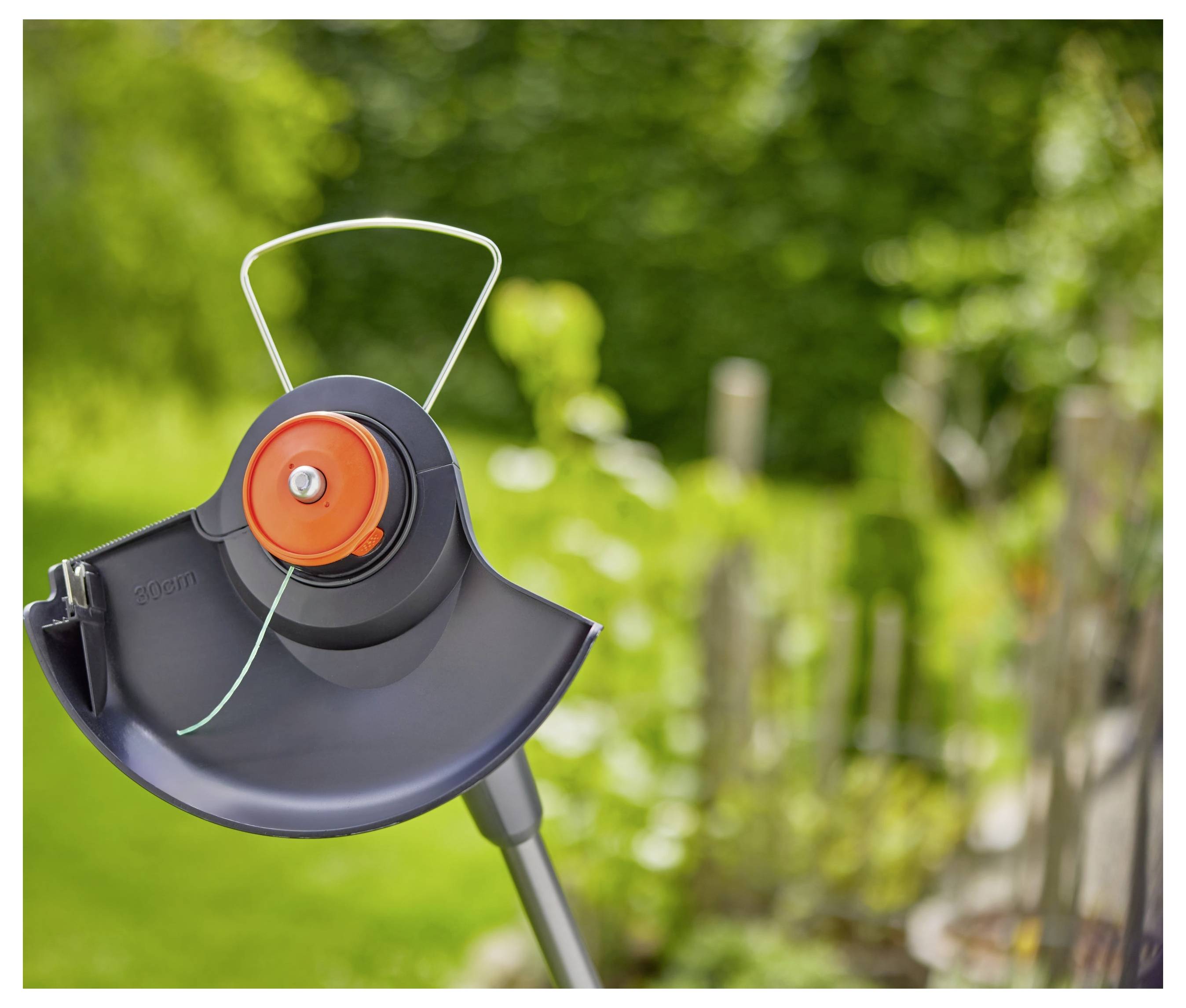 Das Bild zeigt eine Nahaufnahme eines Fadenkopfes eines Motorsensen mit Gras im Hintergrund, wobei die Schneidschnur und der Schutzschild hervorgehoben werden.