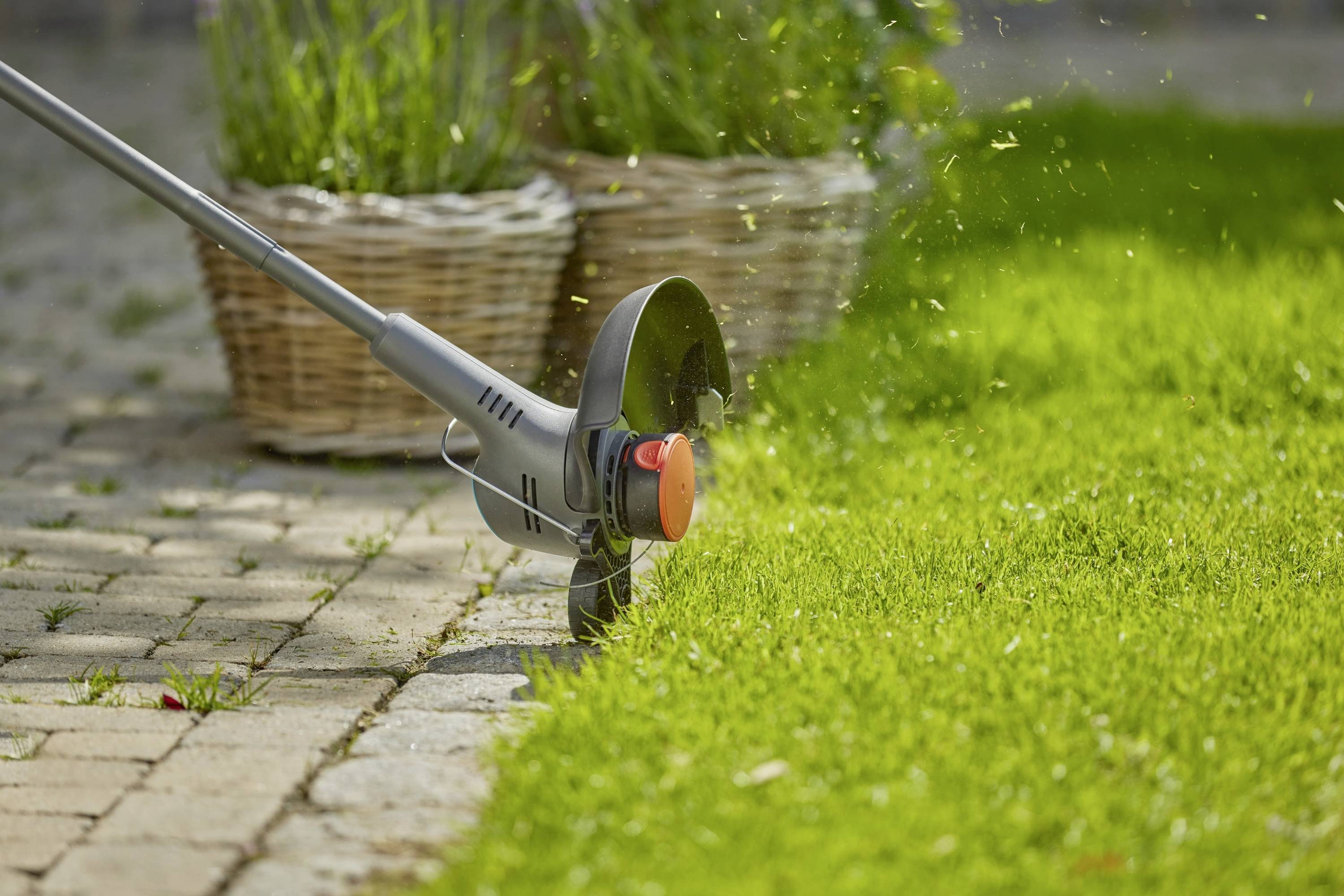 Elektrischer Rasentrimmer, der an einem Steinweg einen Rasenrand schneidet, wobei Grasschnipsel sichtbar durch die Luft fliegen und Gartenunterhaltung veranschaulichen.