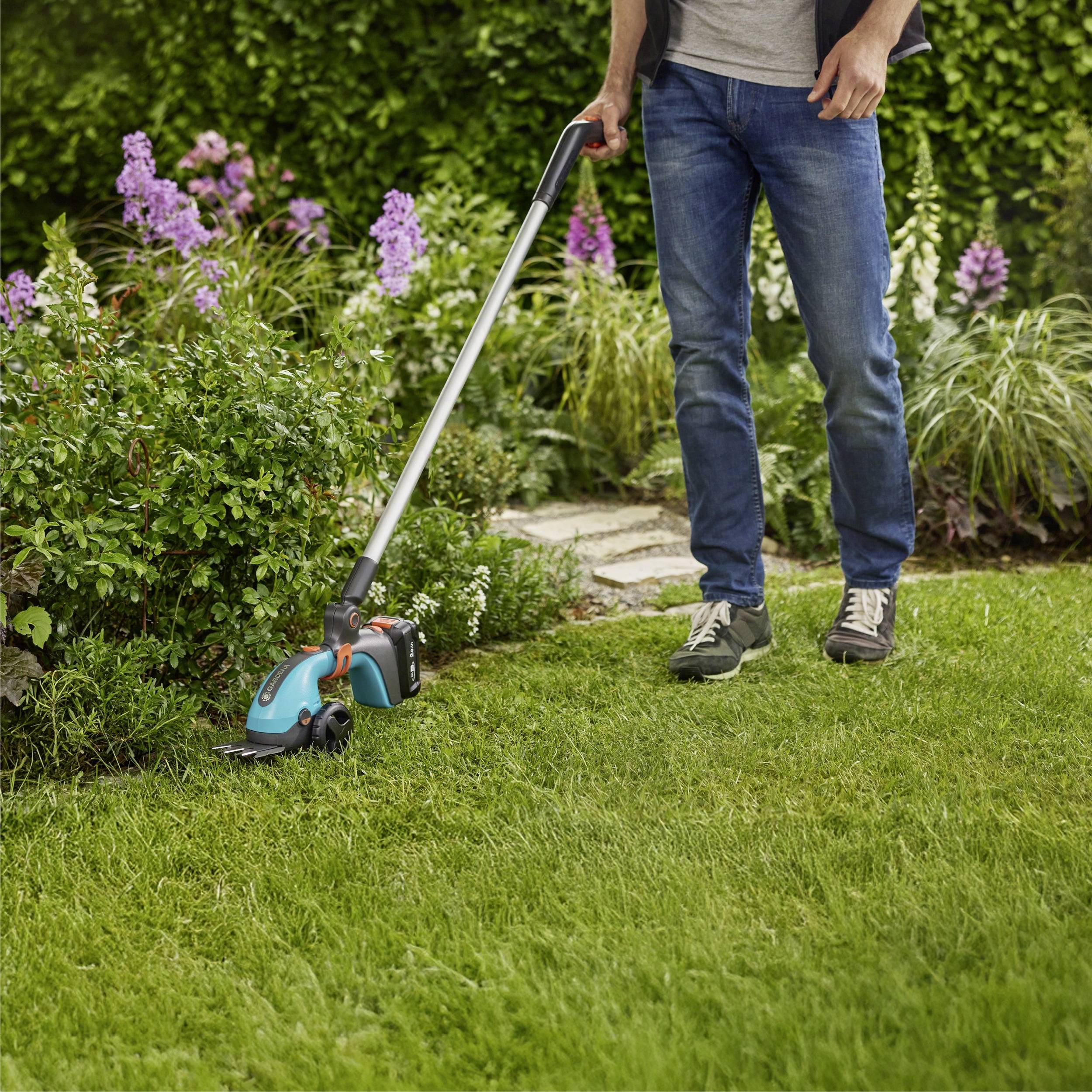 Eine Person trimmt einen Gartenrand mit einem blauen und schwarzen elektrischen Trimmer, umgeben von grünen Büschen und lila Blumen.