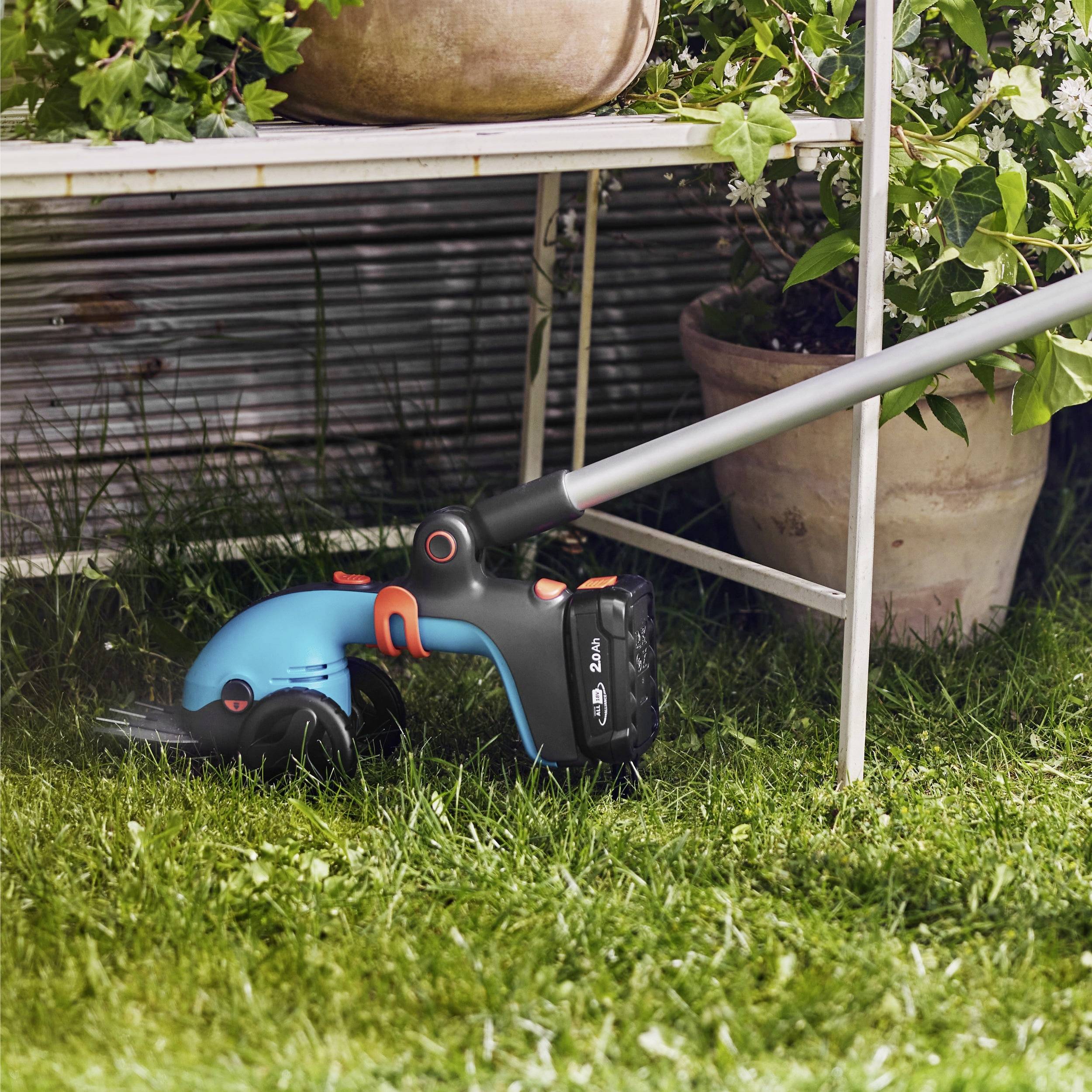 Ein blauer und schwarzer schnurloser Rasentrimmer sitzt auf grünem Gras neben einer Topfpflanze, mit einem Zaun im Hintergrund.
