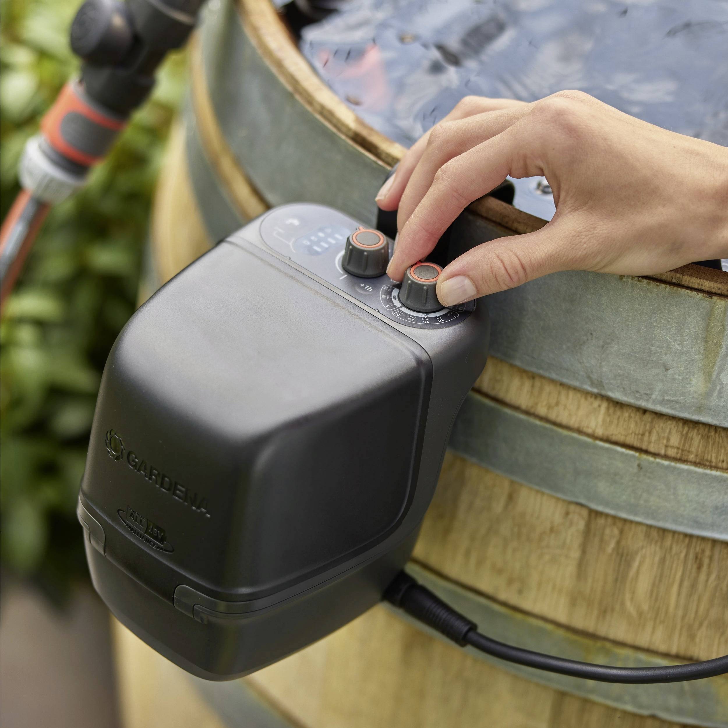 Eine Hand justiert Einstellungen an einem digitalen Wassertimer, der an einem Fass befestigt und mit einem Schlauch verbunden ist, was auf eine Bewässerungs- oder Wasserzuführungsanlage hindeutet.