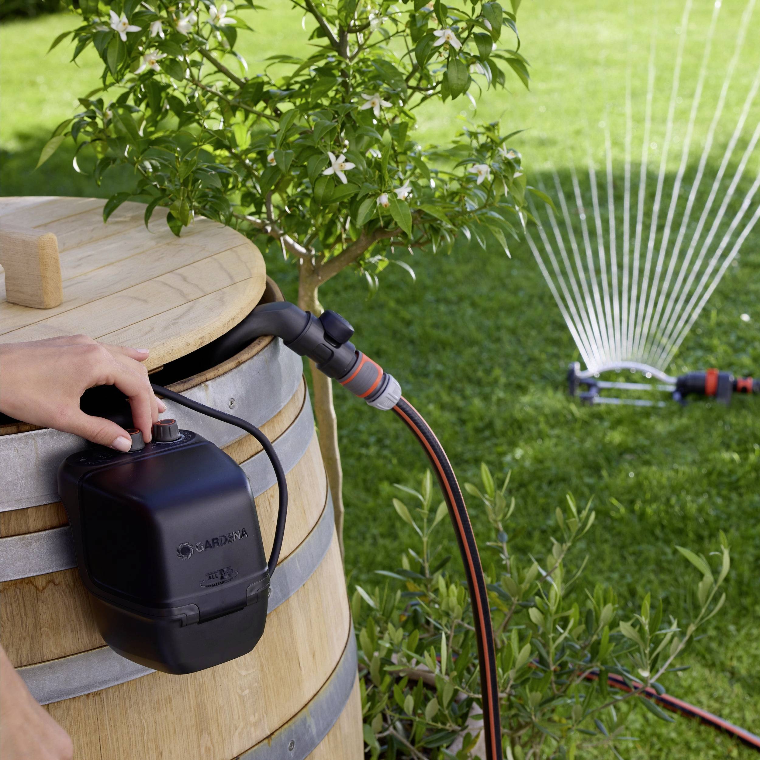 Eine Person stellt den Hahn an einer Holzregentonne ein, die mit einem Schlauch verbunden ist, während ein Sprinkler im Hintergrund einen Rasen bewässert.