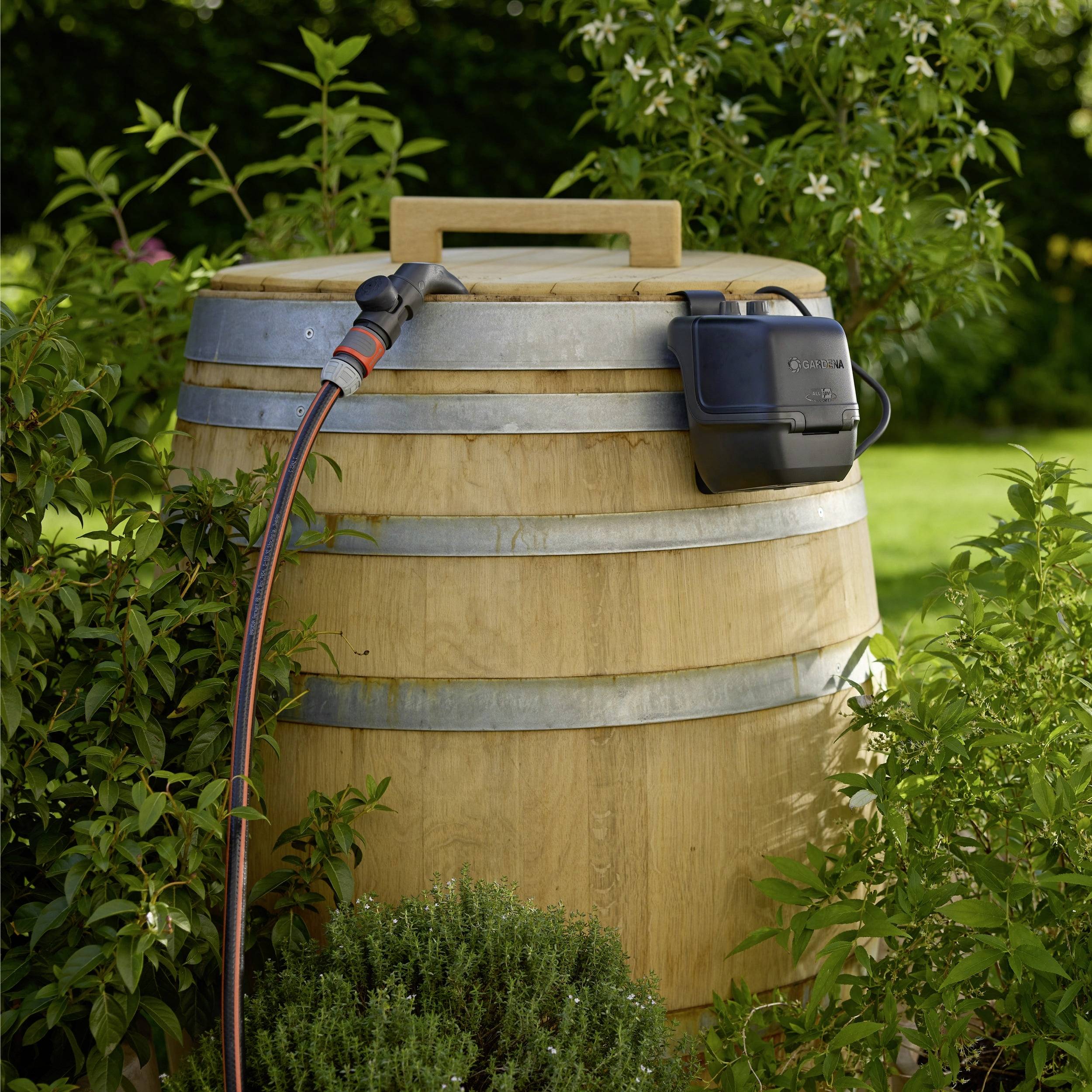 Eine Holzregentonne mit einem angeschlossenen Schlauch steht in einem Garten, umgeben von Grünpflanzen, und demonstriert eine Methode zur Wassersammlung.
