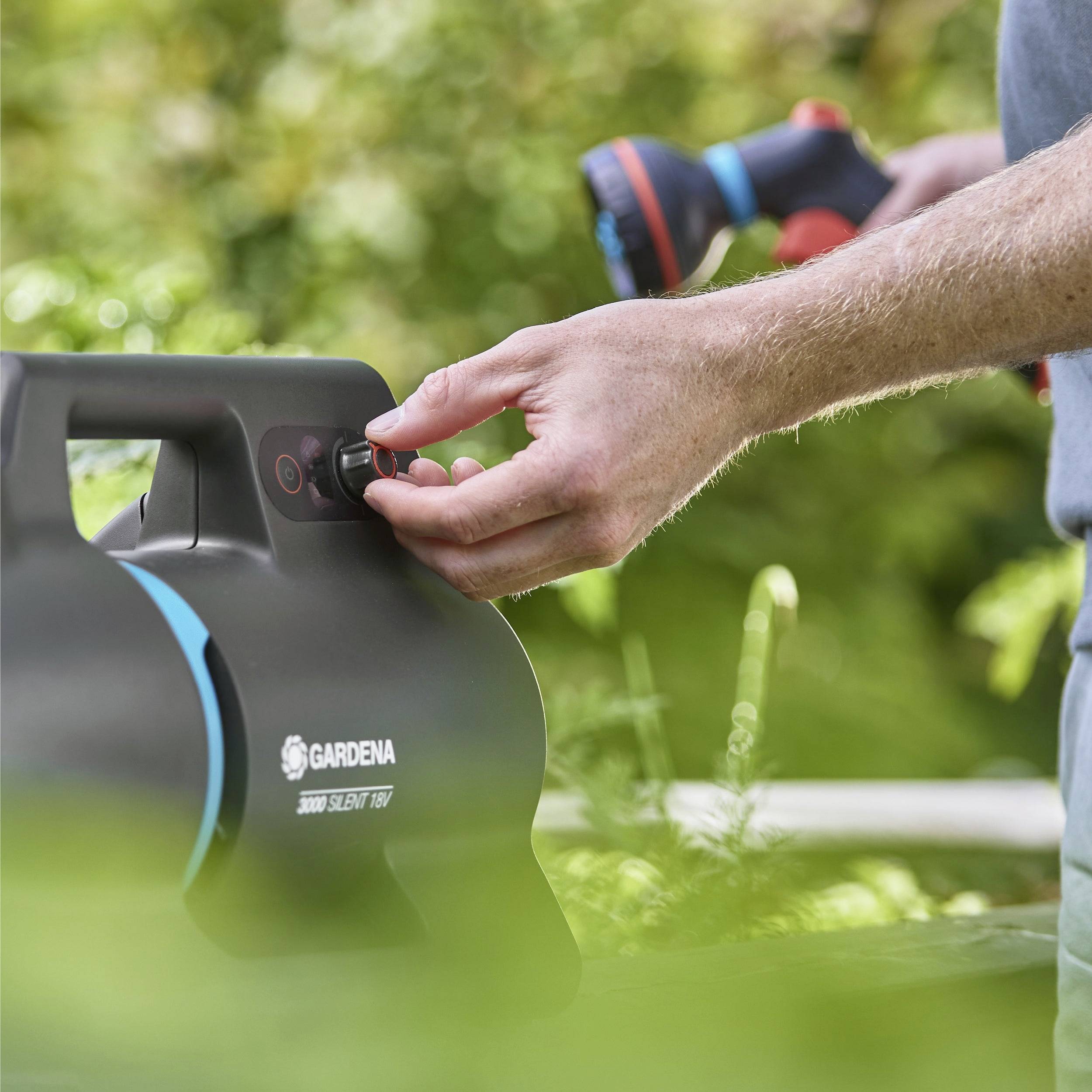 Eine Person, die die Einstellungen an einem 'Gardena 3500 Silent 100' Outdoor-Gerät anpasst, mit Grünpflanzen im Hintergrund, was auf eine Gartenumgebung hindeutet.