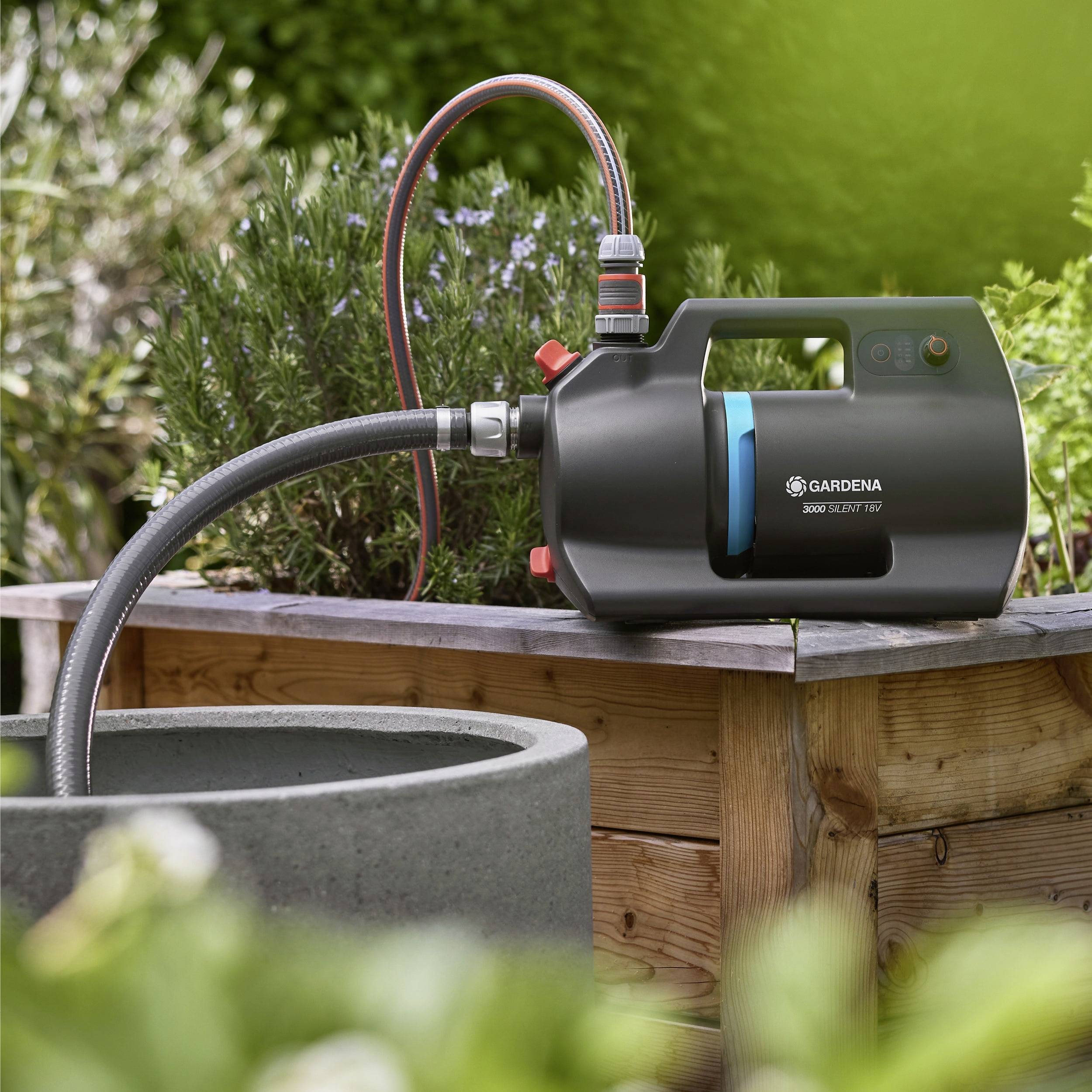 Eine schwarze Wasserpumpe mit roten und grauen Akzenten ist an einen Gartenschlauch angeschlossen und steht auf einer Holzplattform neben einem Pflanzgefäß.