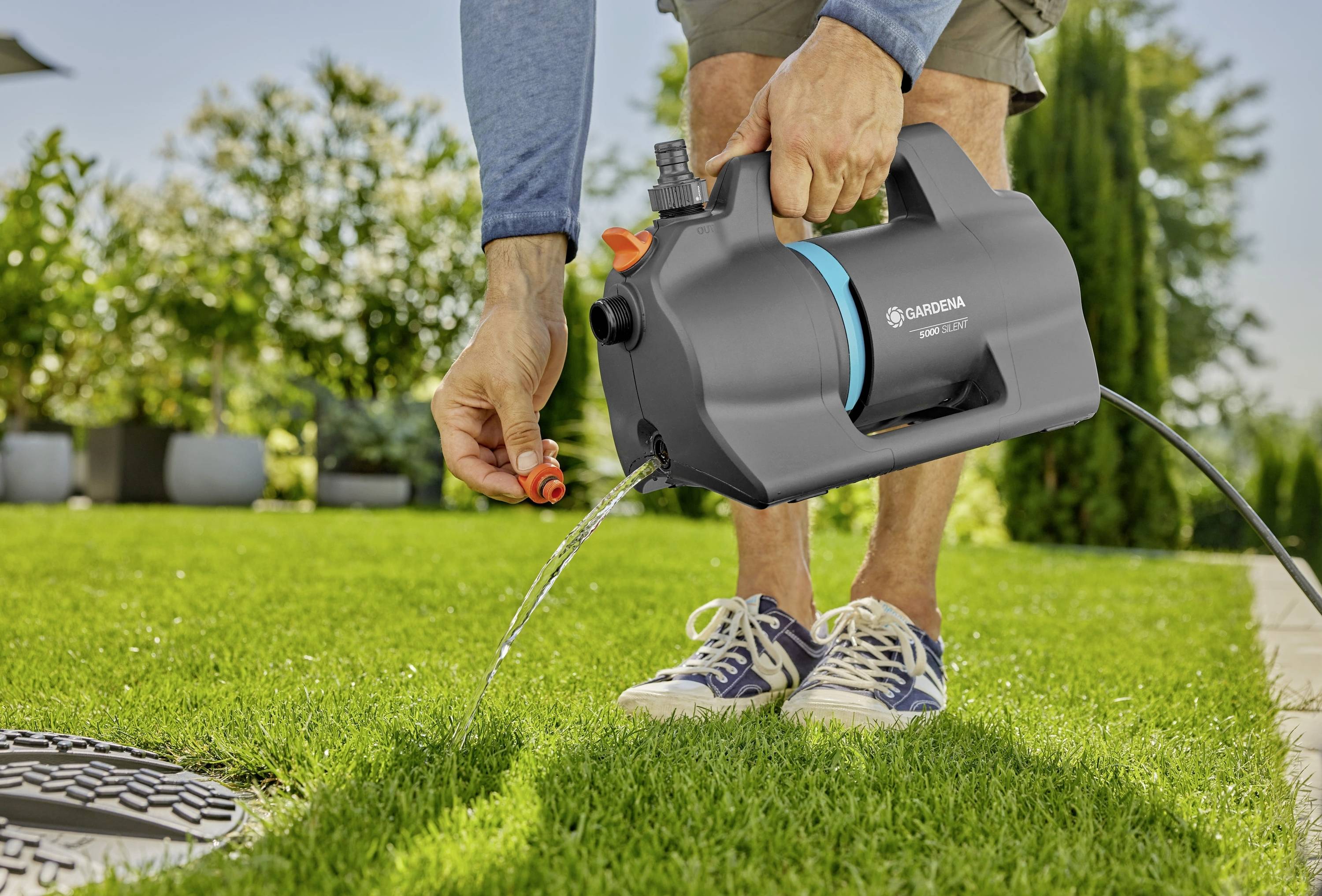 Eine Person bewässert einen Rasen mit einer tragbaren Gardena-Gartenpumpe, wobei Wasser auf das Gras strömt. Sonnendurchfluteter Hintergrund mit Pflanzen.
