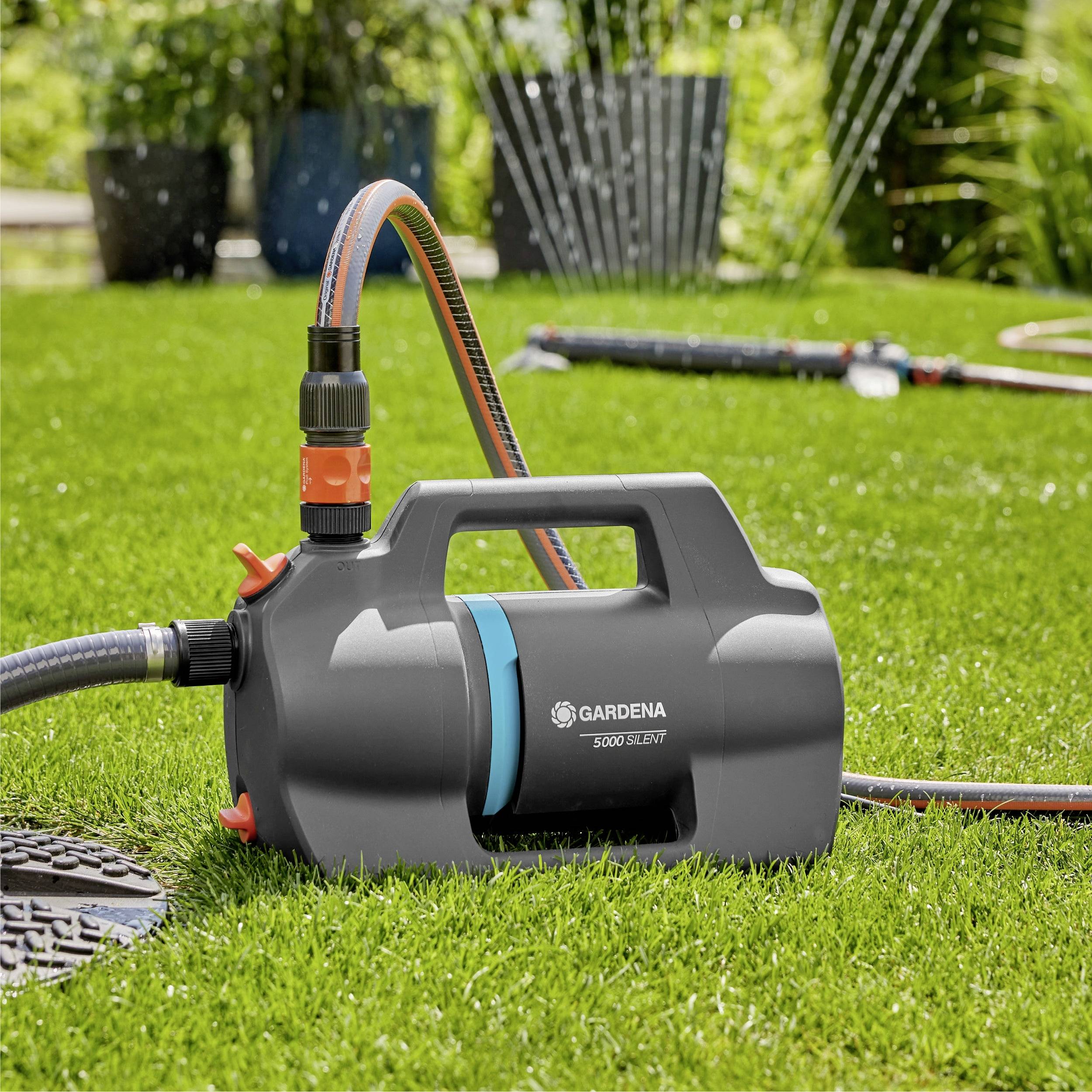 Eine tragbare Gartenpumpe auf Gras mit Sprinklern im Hintergrund. Die Pumpe ist mit der Marke 'Gardena 5000/5E' gekennzeichnet.