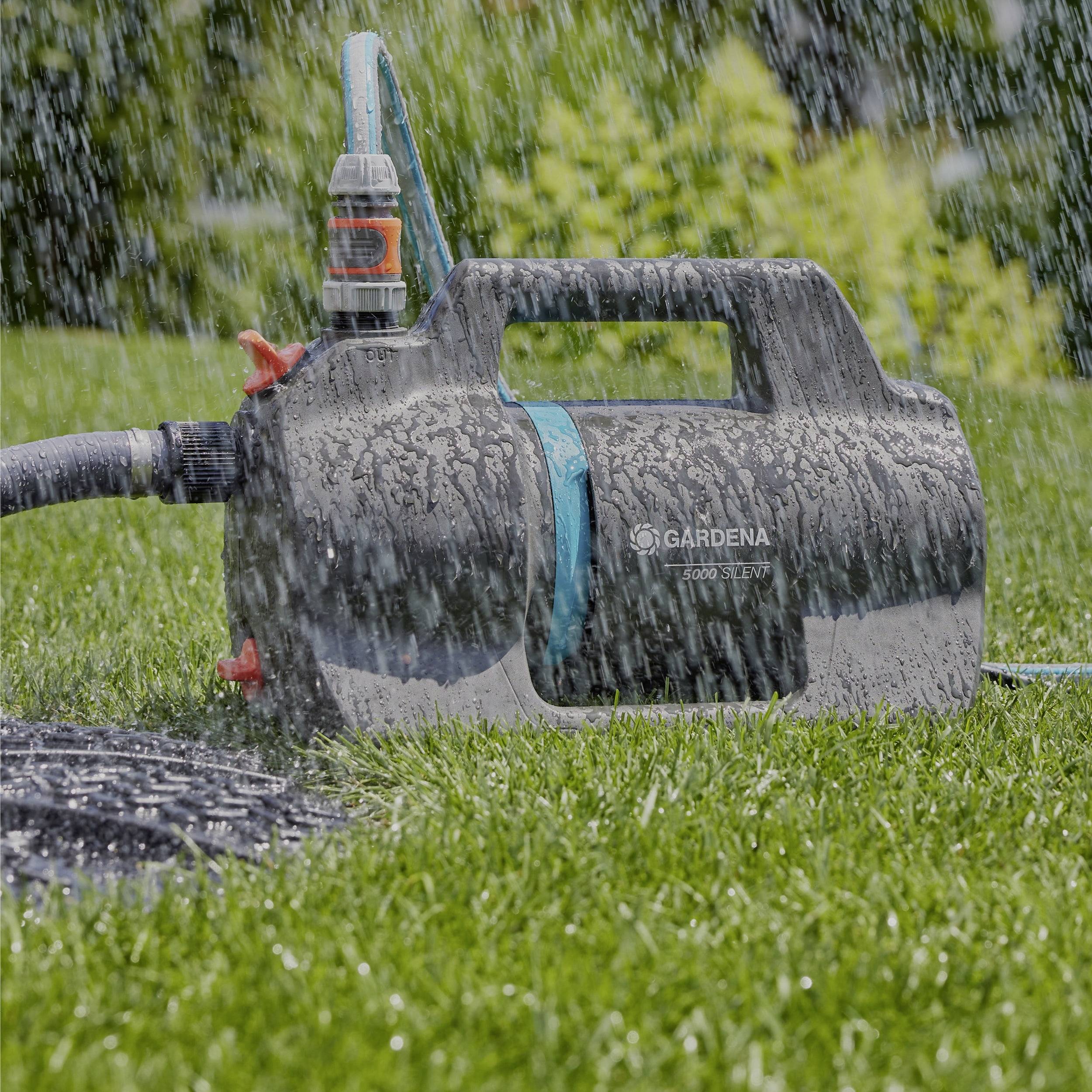 Eine schwarze und blaue elektrische Gartenwasserpumpe versprüht Wasser über einen grasbedeckten Rasen, während Bäume im Hintergrund unscharf erscheinen.