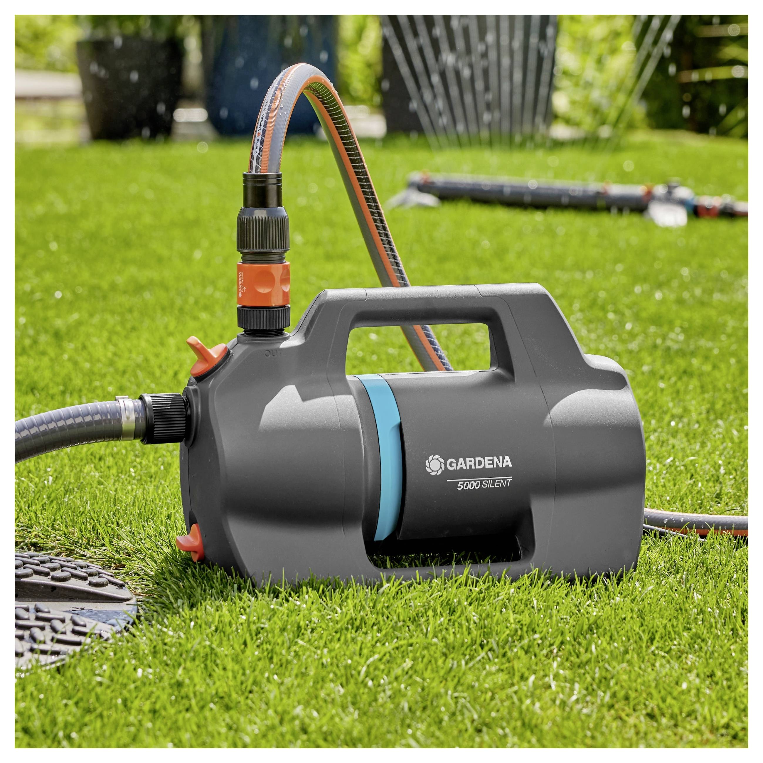 Eine Gardena 3000 Silent Gartenpumpe auf einem Rasen mit zwei angeschlossenen Schläuchen, umgeben von Gartengeräten und Sprinklern im Hintergrund.