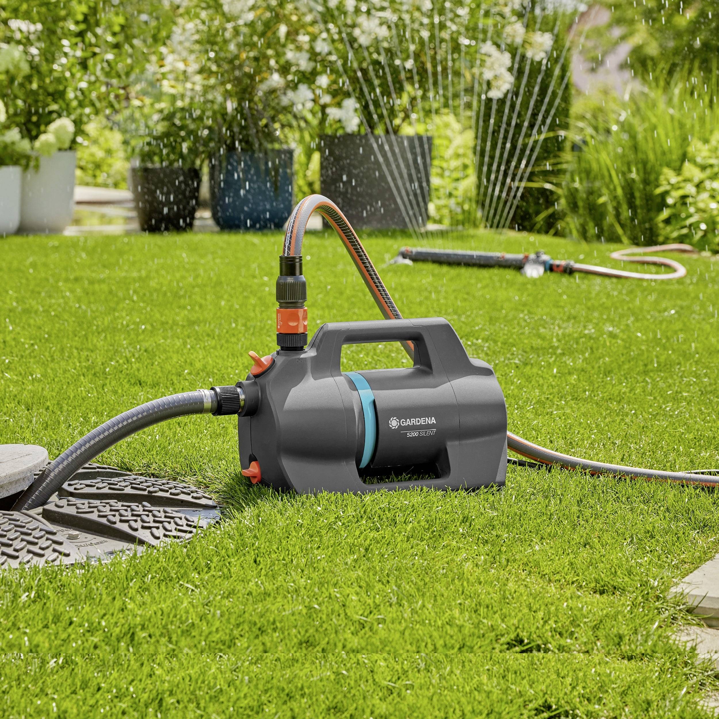 Eine tragbare Wasserpumpe, die auf einem Rasen platziert ist, mit angeschlossenem Schlauch, die zur Gartenbewässerung verwendet wird, umgeben von Pflanzen und einem Sprühstrahl im Hintergrund.