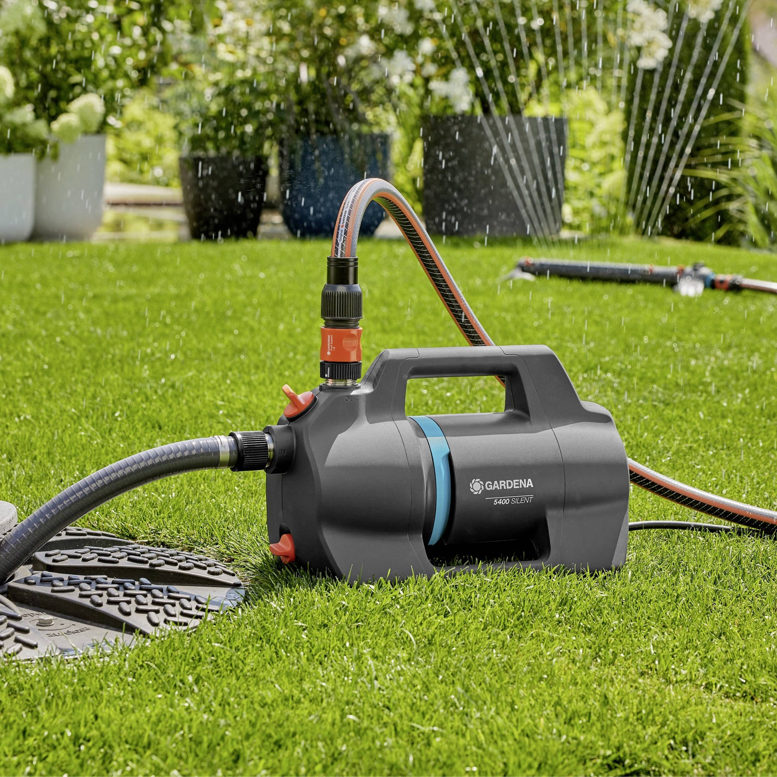 Eine Gartenpumpe steht auf grünem Gras mit angeschlossenen Schläuchen und bewässert aktiv den Rasen und die umliegenden Pflanzen.