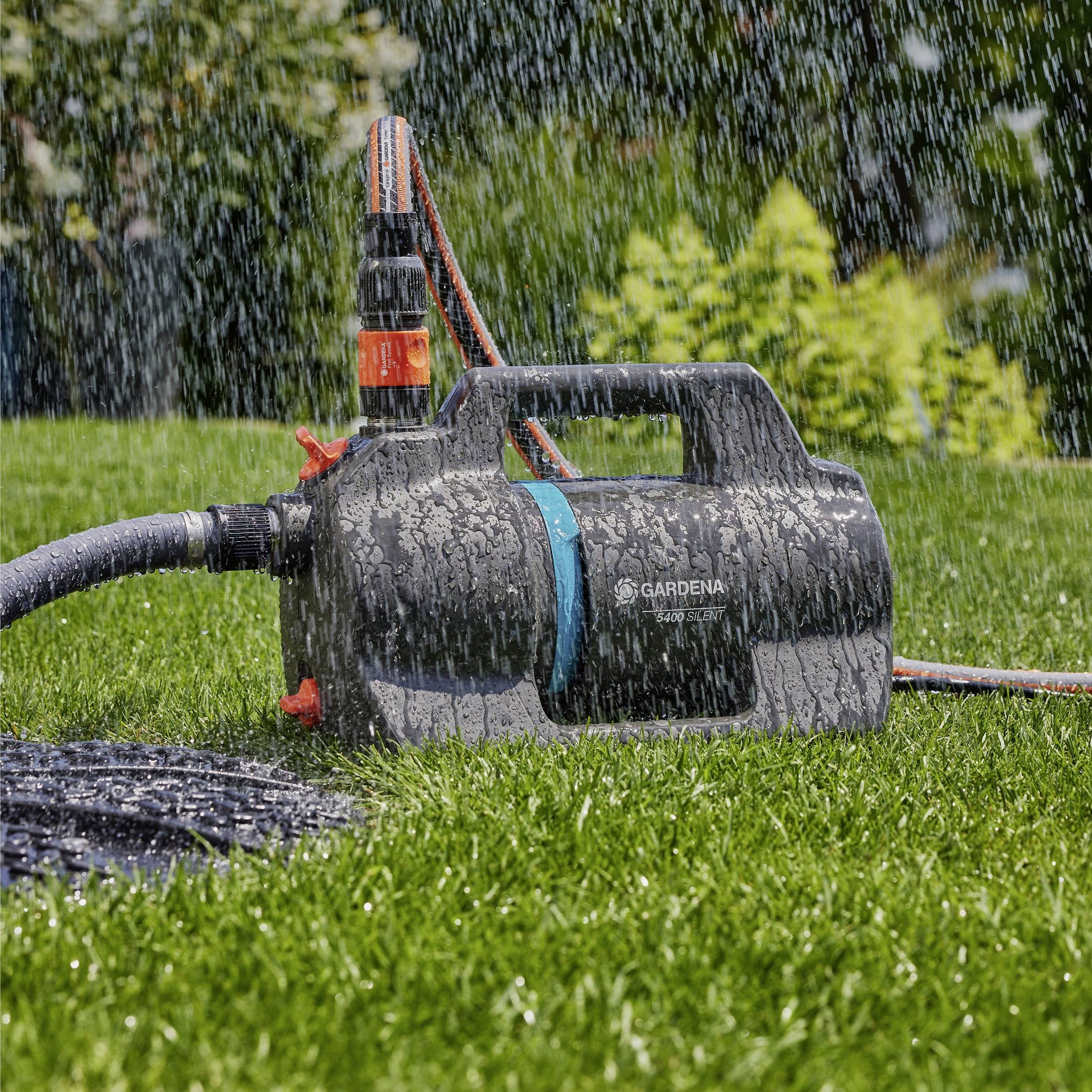 Eine Wasserpumpe steht auf einem Rasen und spritzt Wasser auf das Gras. An der Pumpe ist ein orange-schwarzer Schlauch angeschlossen, während im Hintergrund Grünpflanzen zu sehen sind.