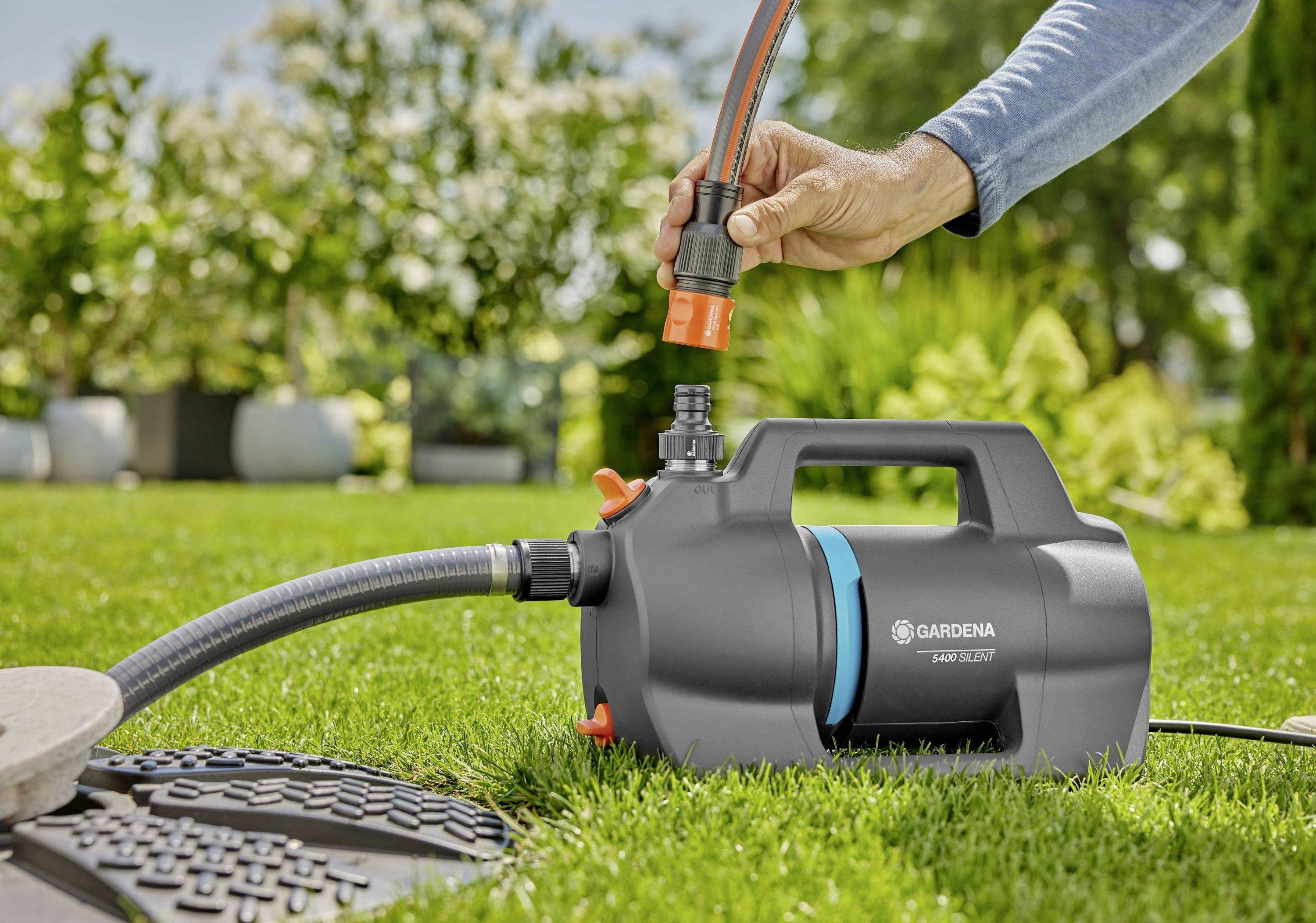 Eine Person verbindet einen Schlauch mit einer Gardena-Gartenpumpe auf einem Rasen, umgeben von Grünflächen und Blumenkübeln in einer sonnigen Außenumgebung.