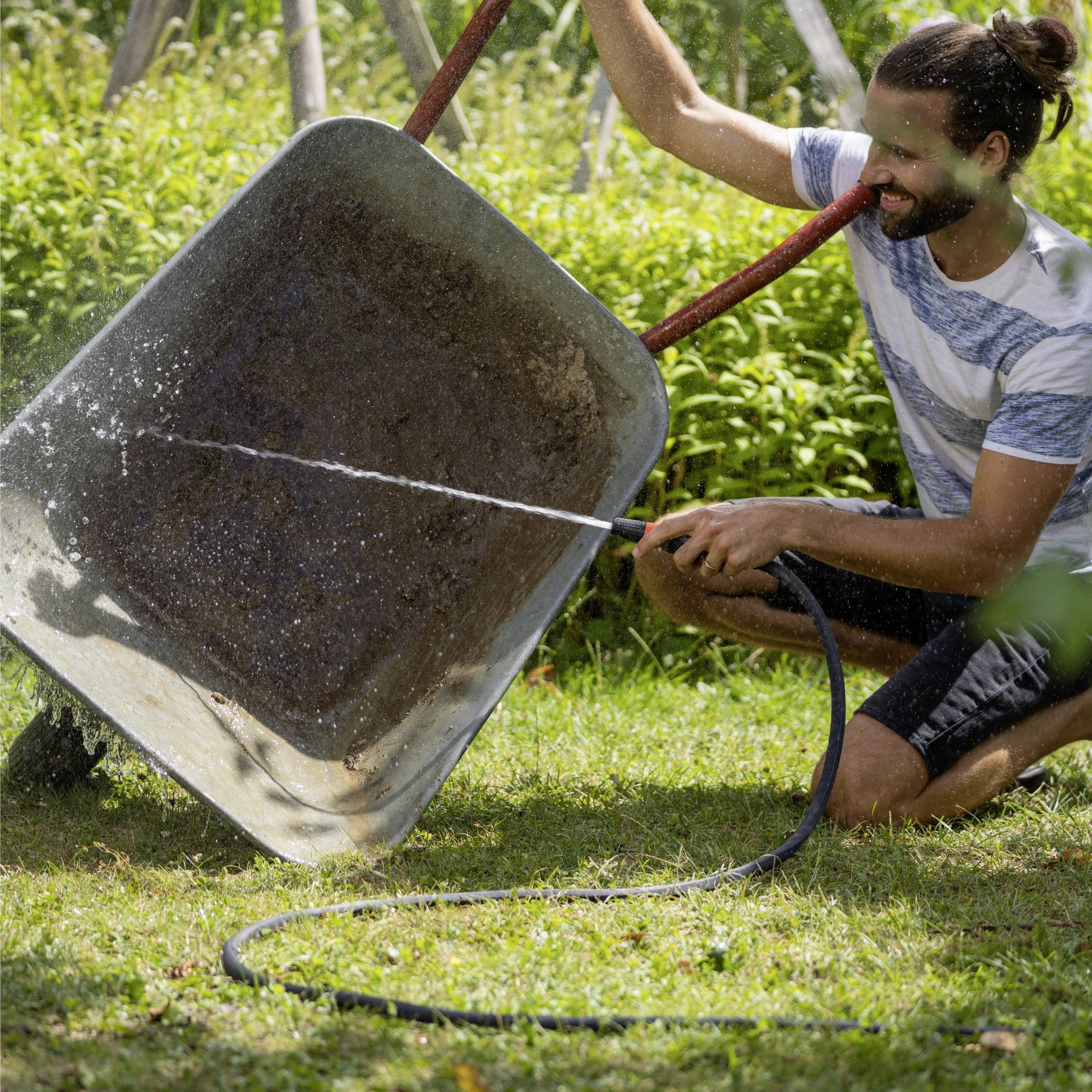 Eine Person lächelt, während sie eine Schubkarre mit einem Gartenschlauch in einem sonnigen Garten umgeben von Grünpflanzen wäscht.