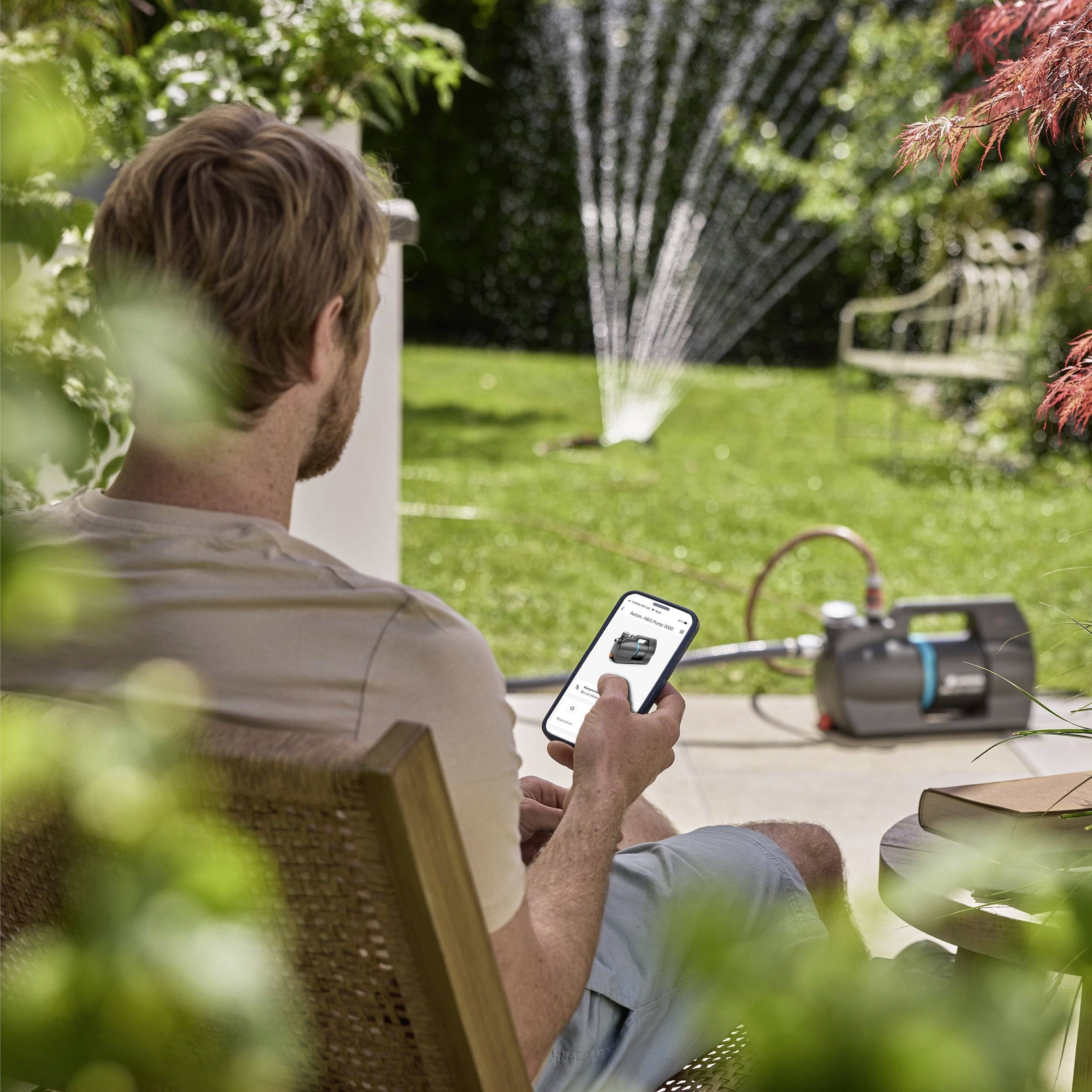 Ein Mann in lässiger Kleidung wäscht eine Schubkarre mit einem Gartenschlauch in einem sonnigen Garten, umgeben von grünem Gras und Pflanzen.