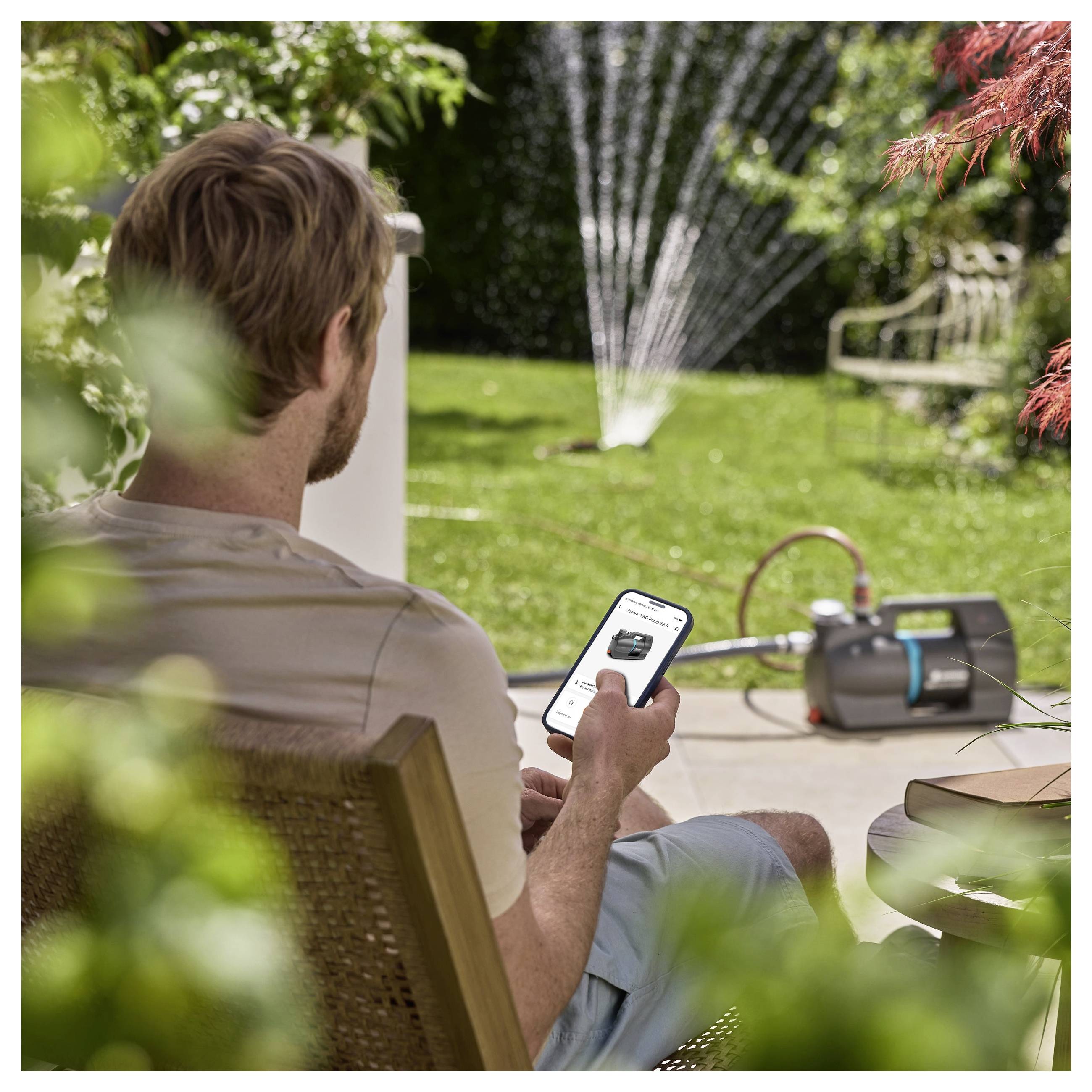 Eine Person, die in einem Garten sitzt, nutzt ein Smartphone, um ein Bewässerungssystem zu steuern und demonstriert dabei eine ferngesteuerte Gartentechnologie.