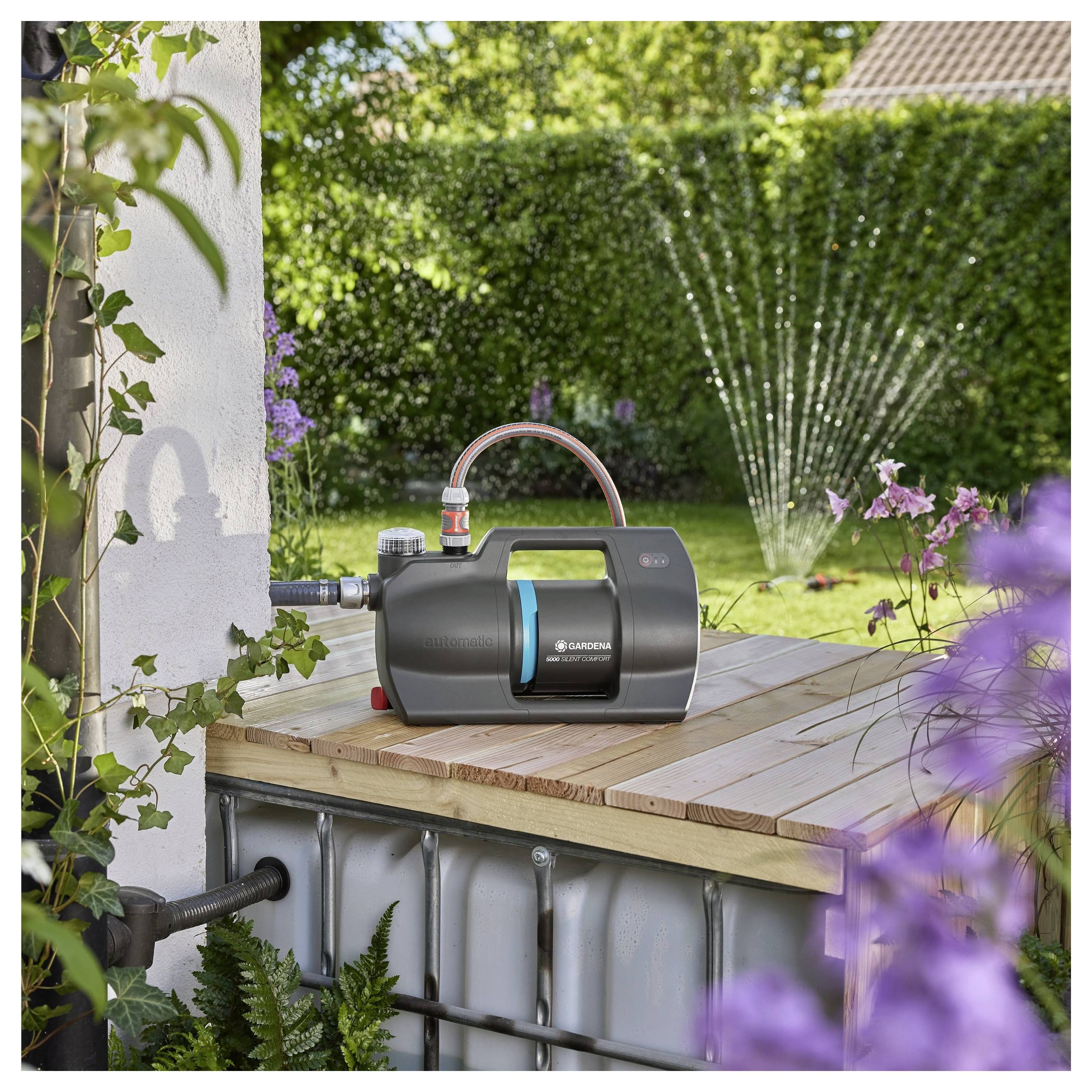 Eine Gartenpumpe steht auf einer Holzplattform in der Nähe eines Wassertanks, während ein Sprinklersystem einen üppig grünen Rasen und blühende Pflanzen im Hintergrund bewässert.