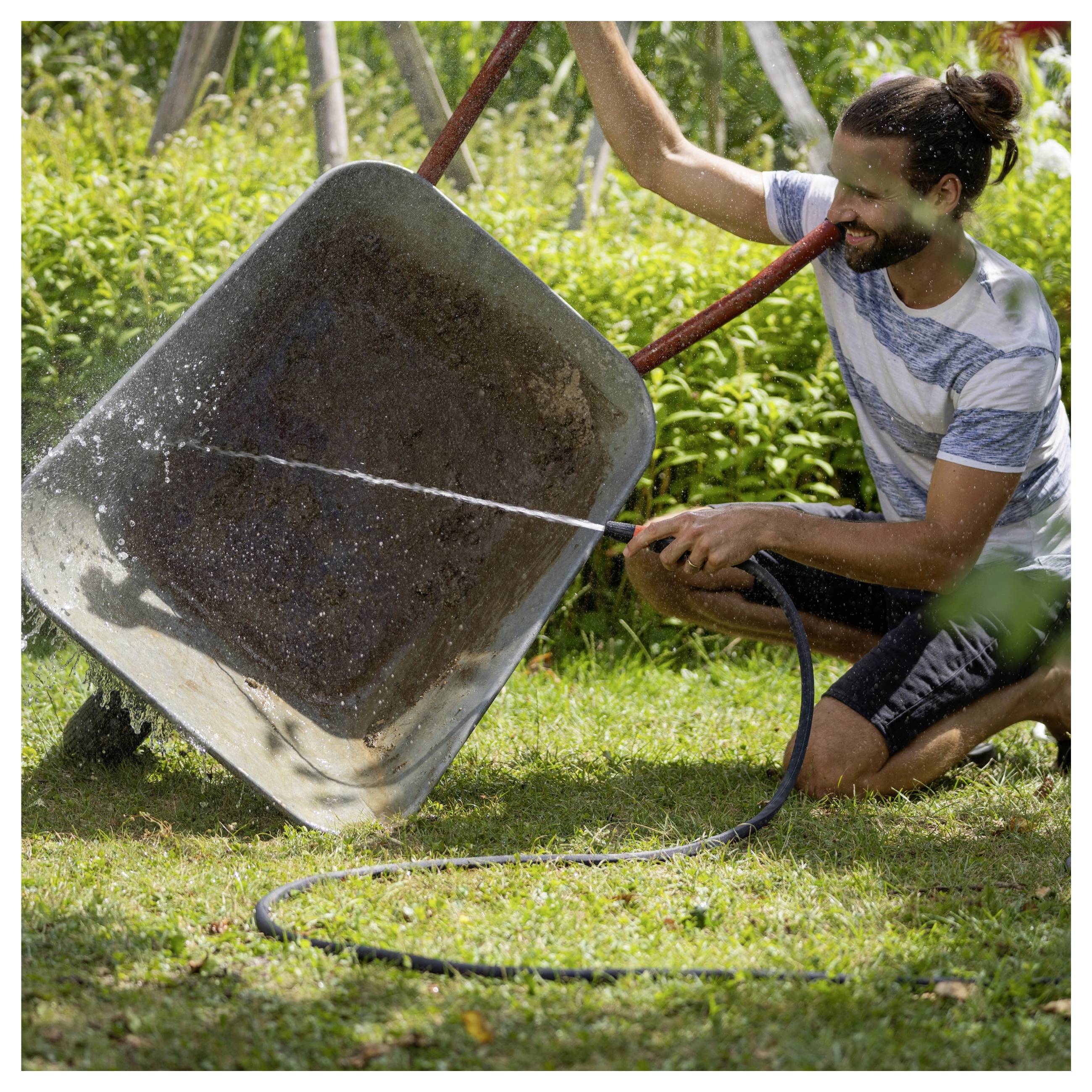 Ein Mann in einem gestreiften Hemd benutzt einen Schlauch, um eine Metallschubkarre zu reinigen, sprüht Wasser darauf, in einer grünen Gartenumgebung.