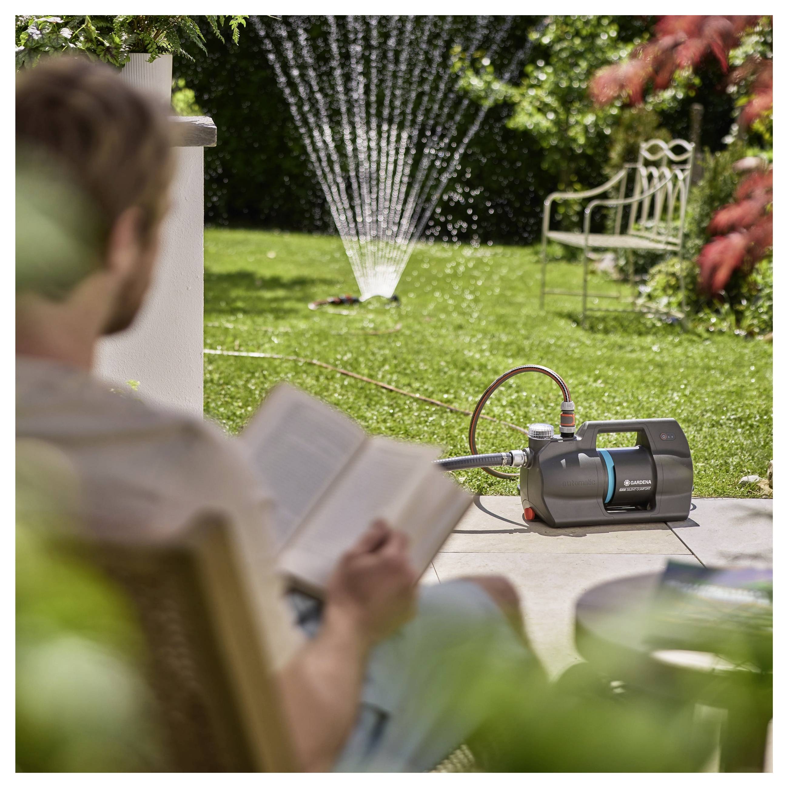 Eine Person liest ein Buch in einem Garten, während ein Sprinkler im Hintergrund den Rasen bewässert und eine entspannende Außenszenerie schafft.