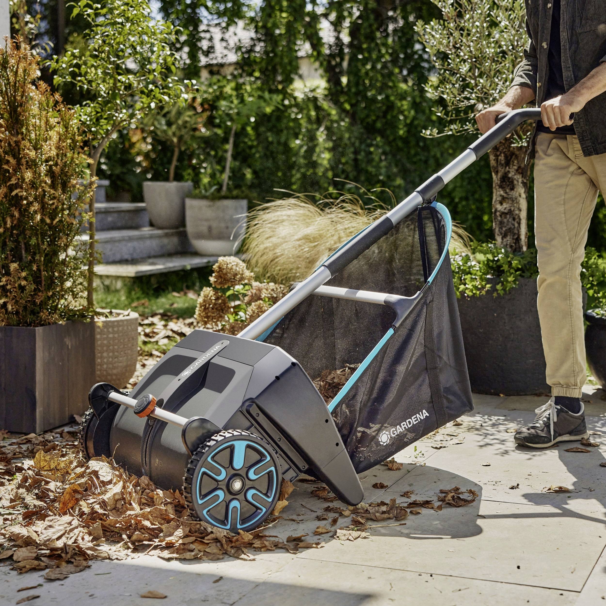 Eine Person, die einen manuell betriebenen Laubsammler verwendet, um gefallene Blätter auf einer Terrasse neben Topfpflanzen bei sonnigem Wetter zu sammeln.
