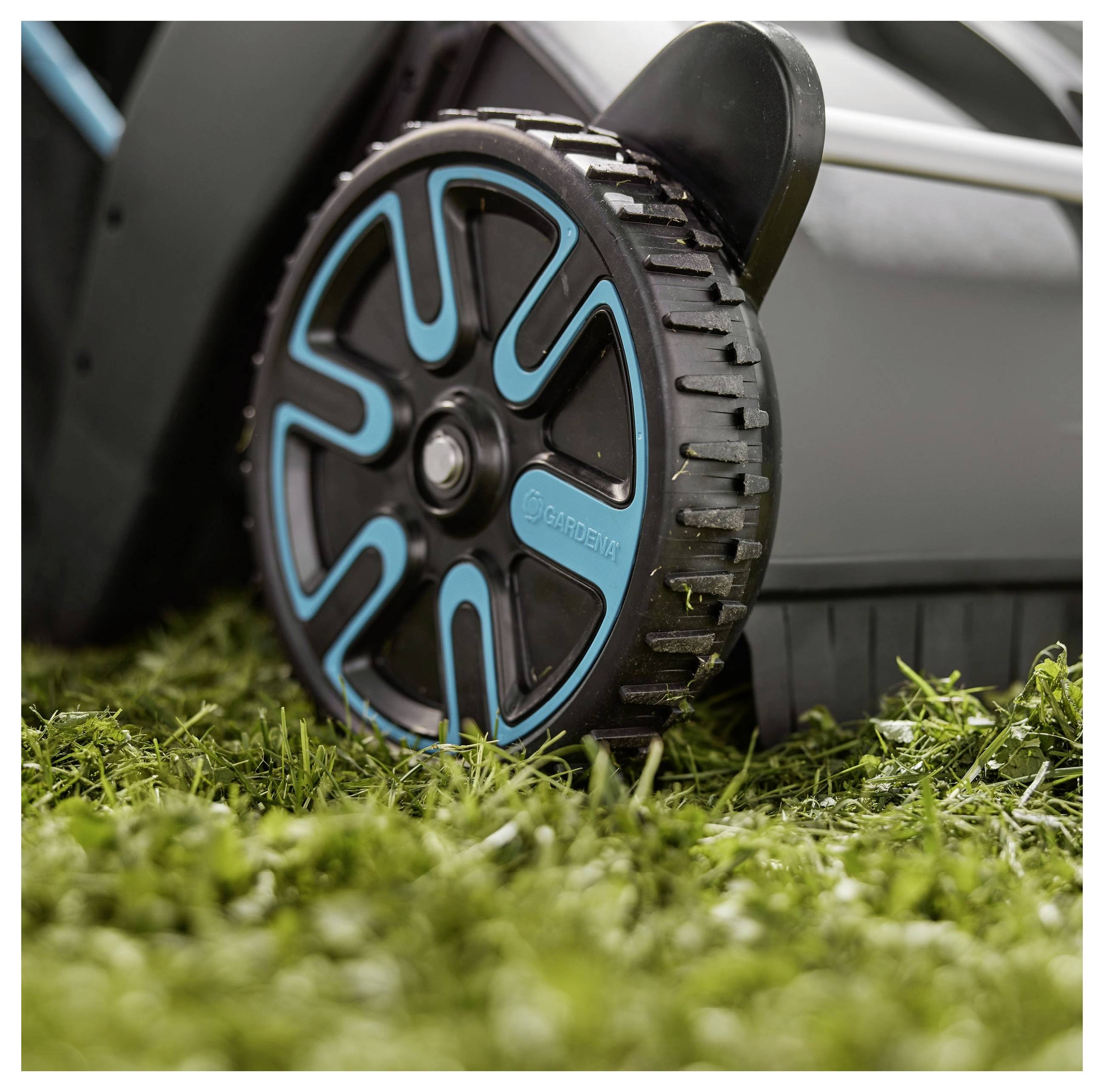 Nahaufnahme eines Rasenmäherades auf Gras. Das Rad hat ein schwarzblaues Design mit sichtbarem Profil und zeigt die Marke „Gardena