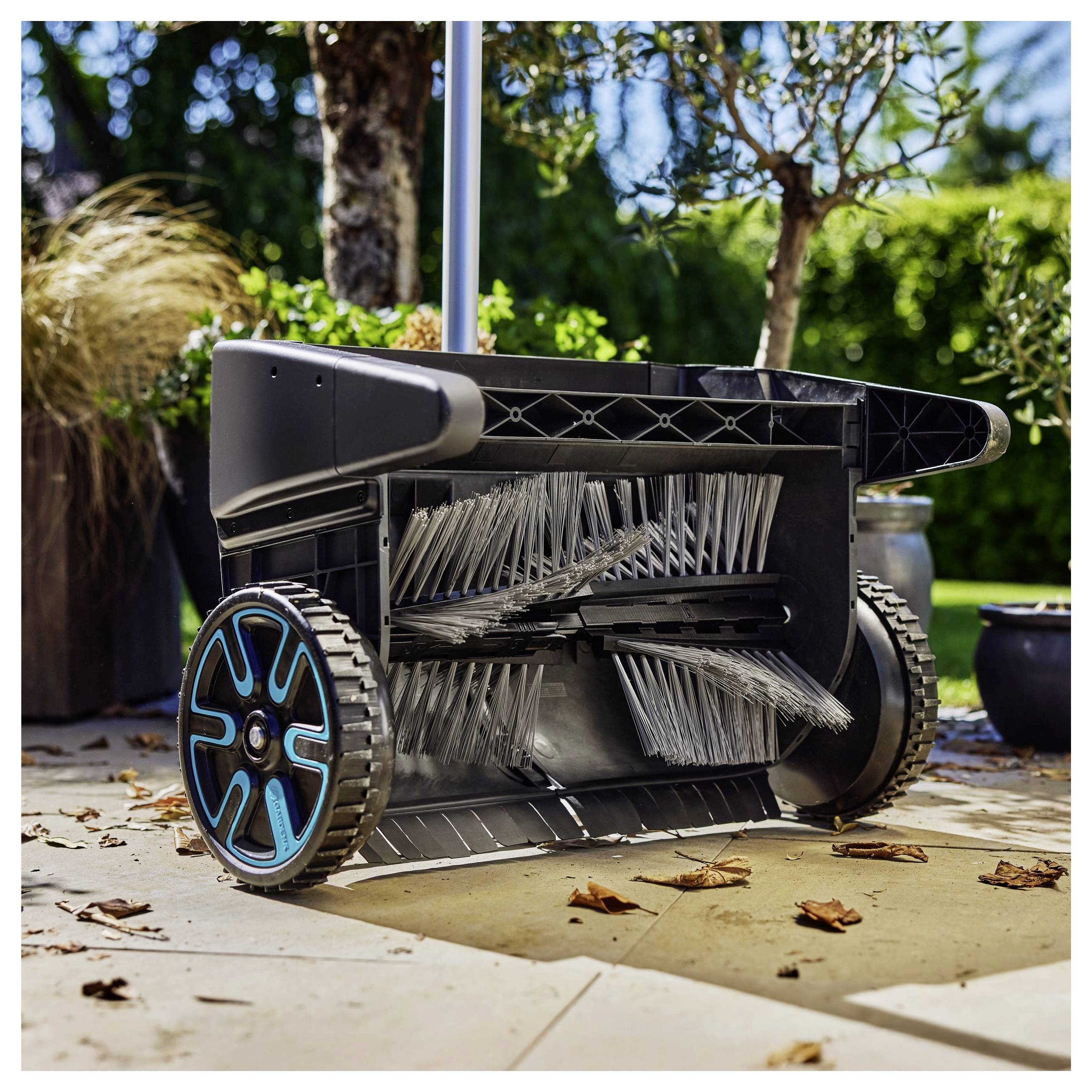 Ein Kehrmaschine mit rotierenden Bürsten und einem Auffangbehälter steht auf einer Steinterrasse, umgeben von Pflanzen in einer Gartenanlage.