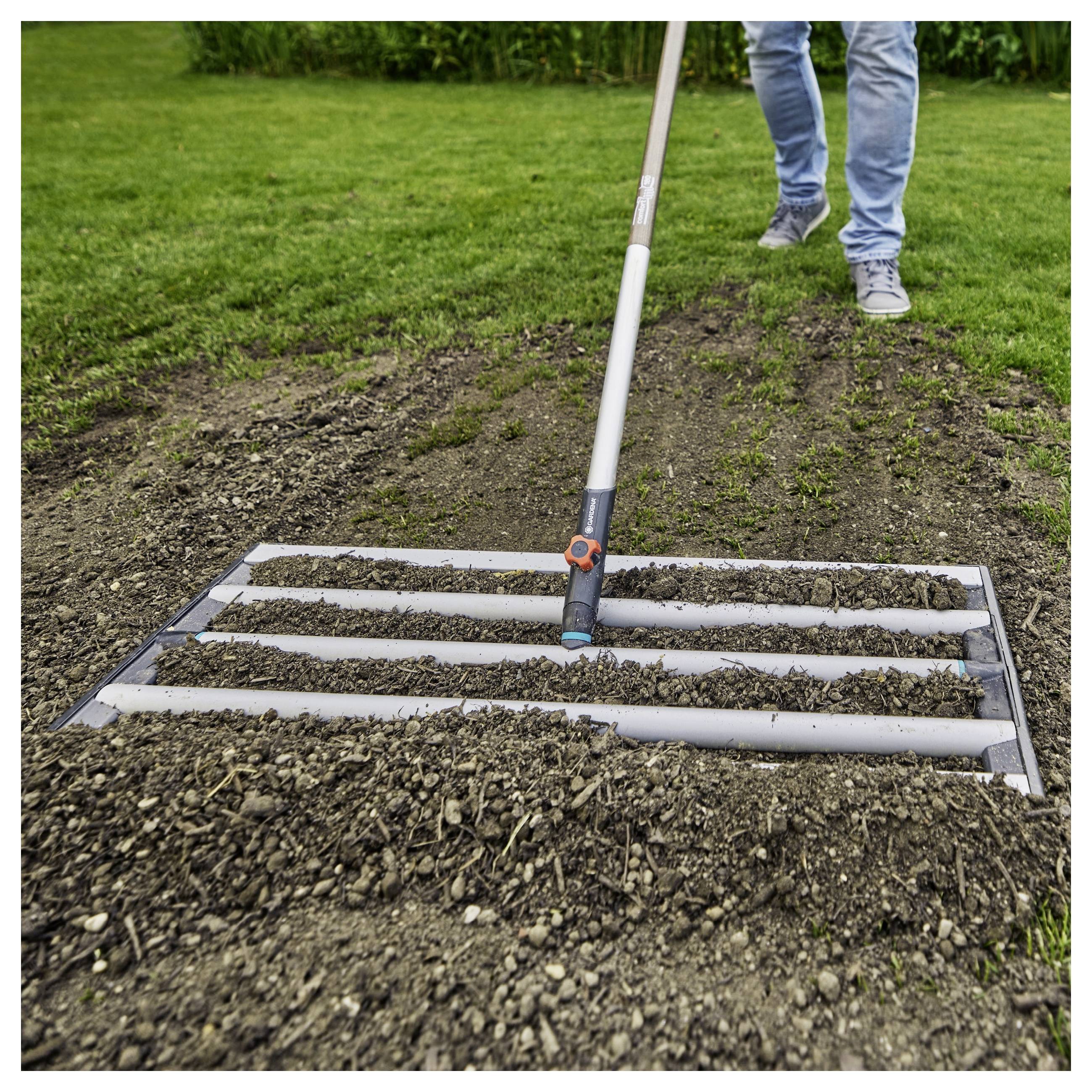 Eine Person benutzt ein Gartenwerkzeug mit horizontalen Stangen, um frisch umgegrabenen Boden auf einem Rasen zu ebnen, was auf eine Vorbereitung zum Pflanzen hindeutet.
