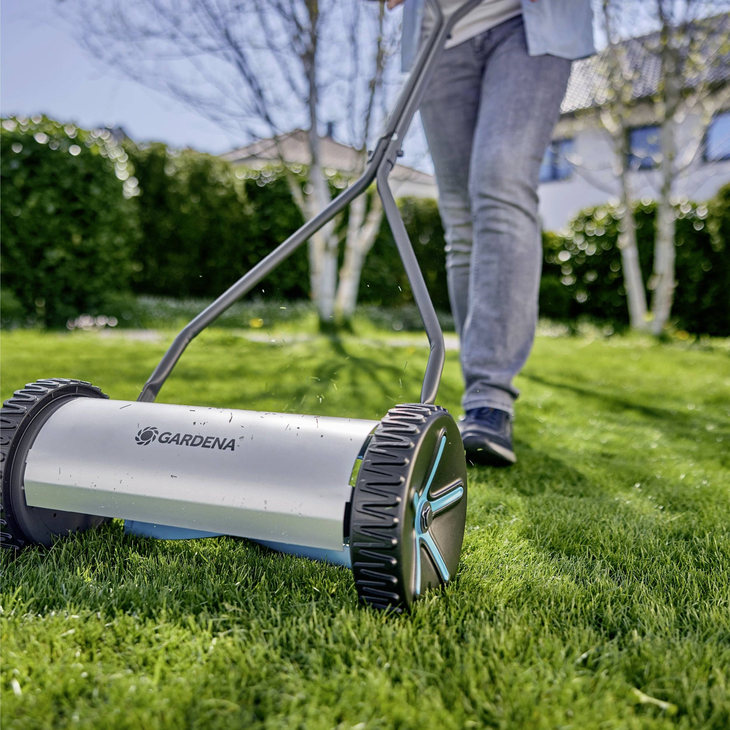 Eine Person, die an einem sonnigen Tag einen manuellen Rasenmäher schiebt und dabei einen Rasen mäht, mit grünem Gras und Bäumen im Hintergrund.