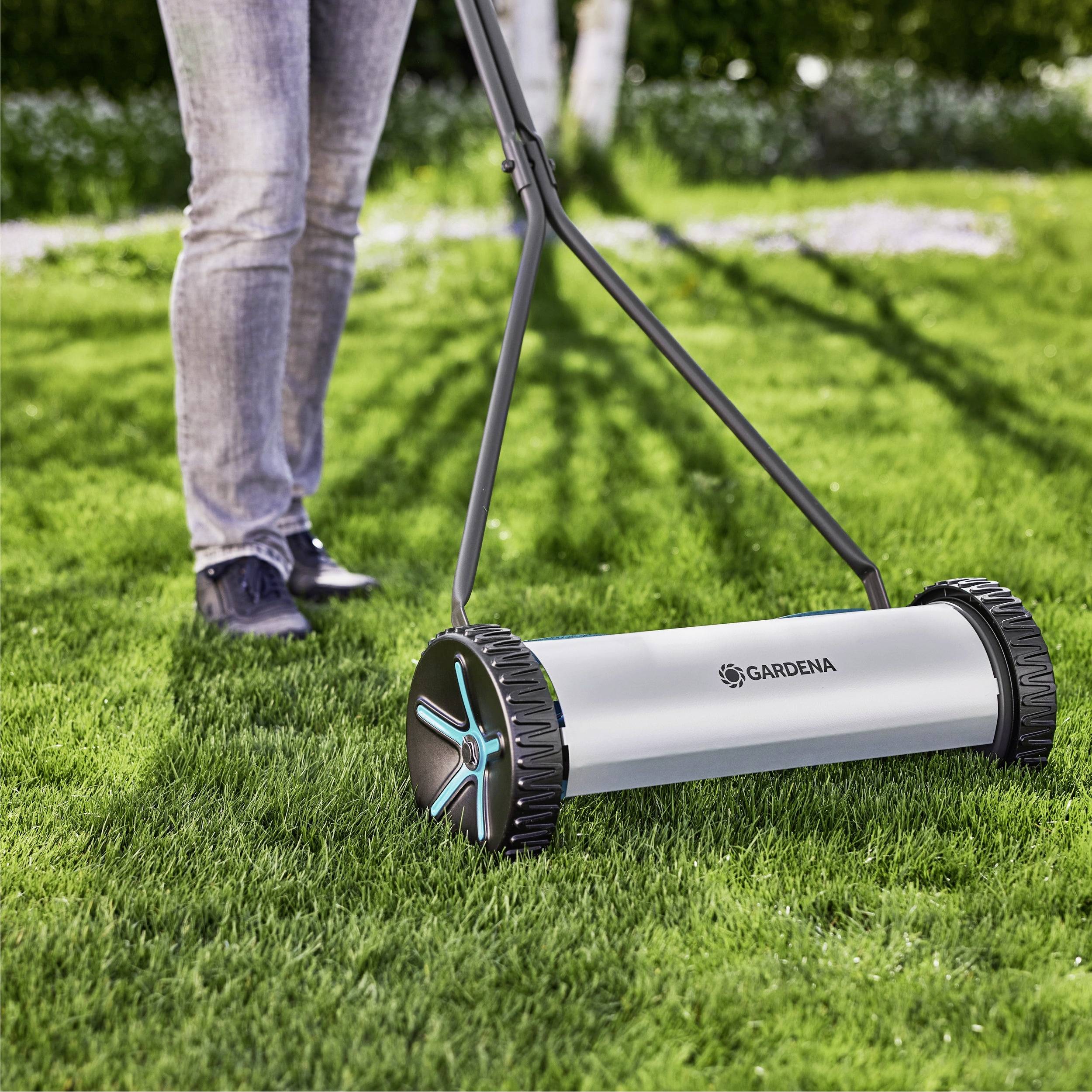 Eine Person, die einen Gardena Rasenmäher schiebt, auf einem grünen Rasen, wobei das Produkt in Aktion gezeigt wird mit Fokus auf sein Design und seine Benutzerfreundlichkeit.
