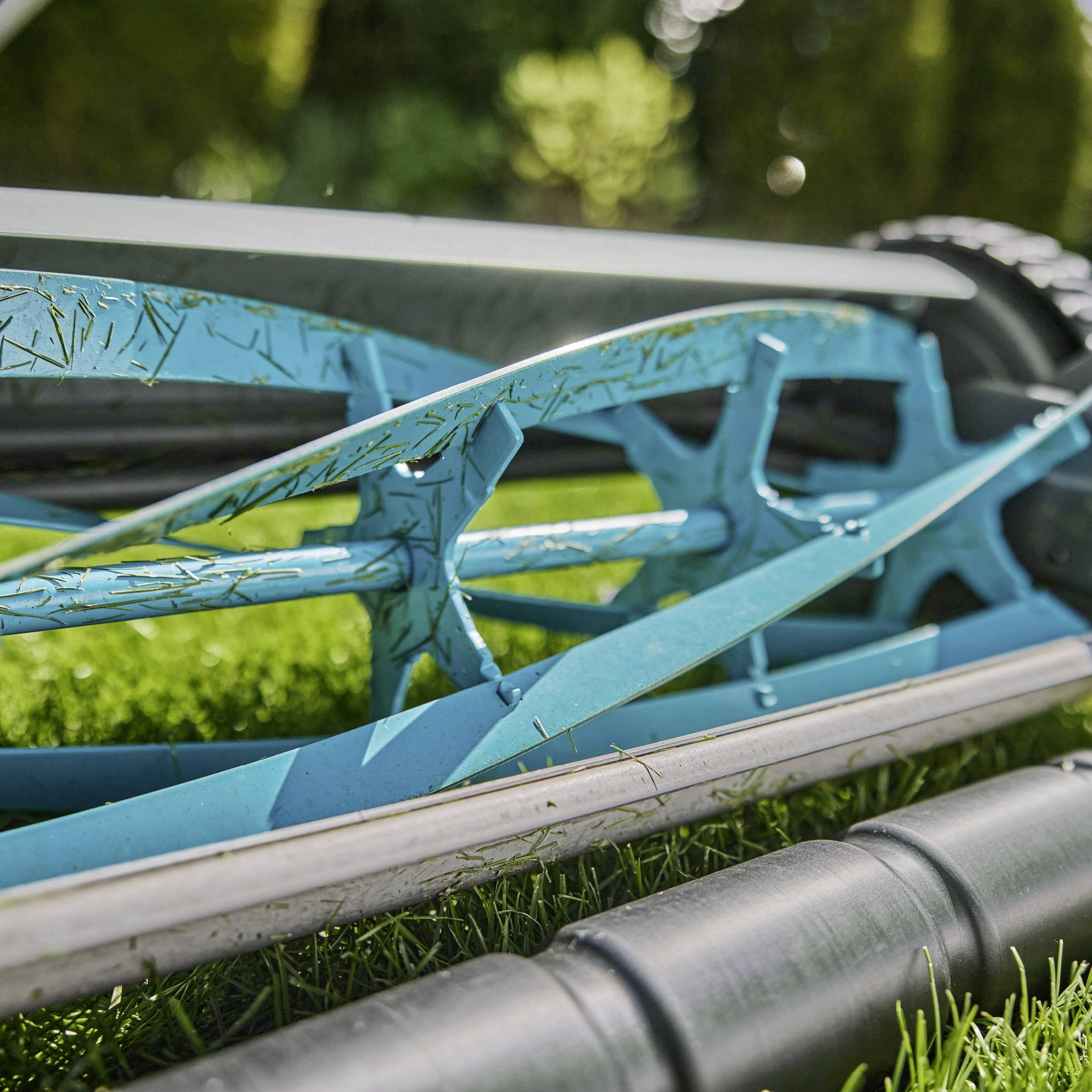 Eine Nahaufnahme eines manuellen Rasenmähers auf Gras, die seinen blauen Metallrahmen, rotierende Klingen und detaillierte mechanische Komponenten zeigt.