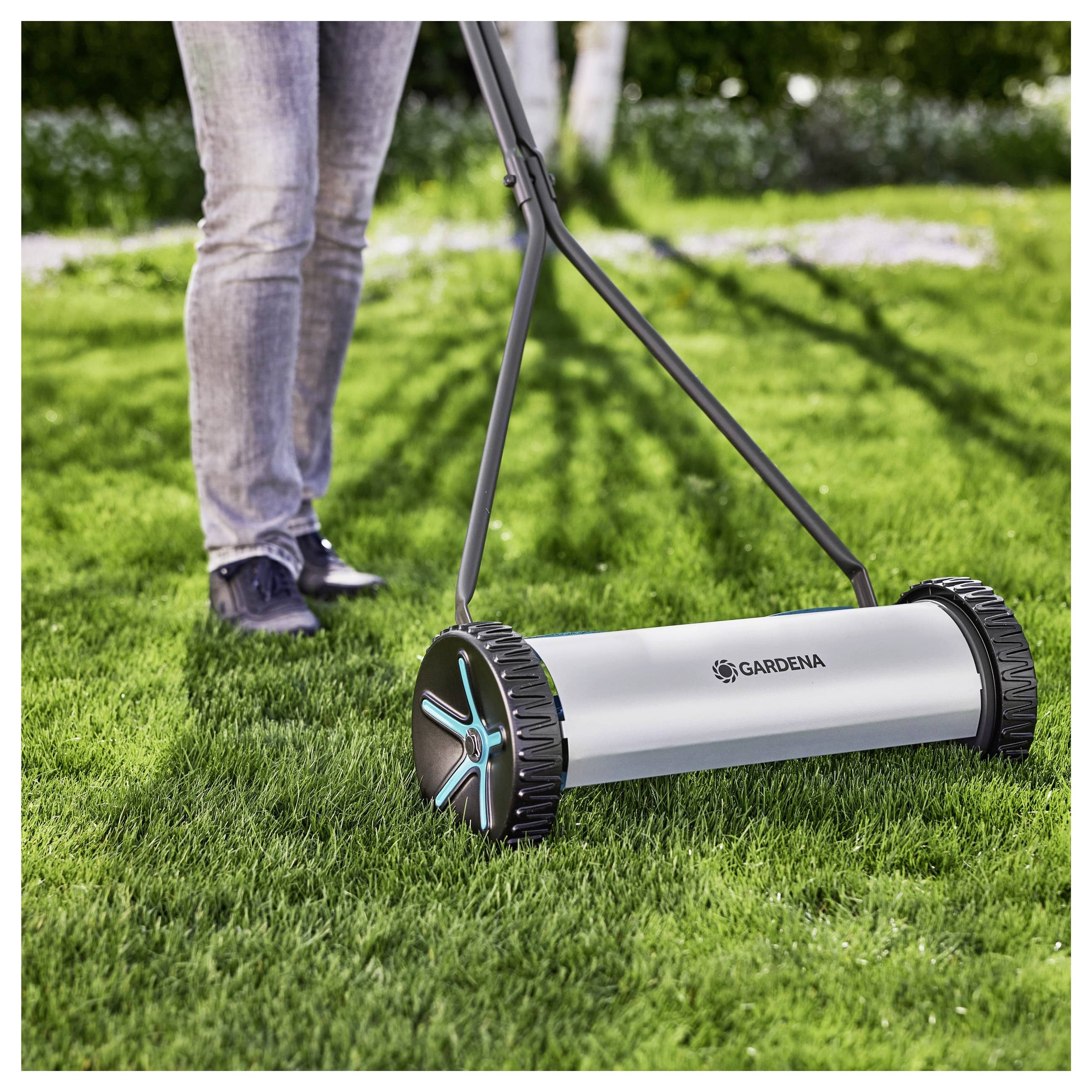 Eine Person mäht einen Rasen mit einem Spindelmäher an einem sonnigen Tag, wobei der Fokus auf dem Design des Mähers und dem üppig grünen Gras liegt.