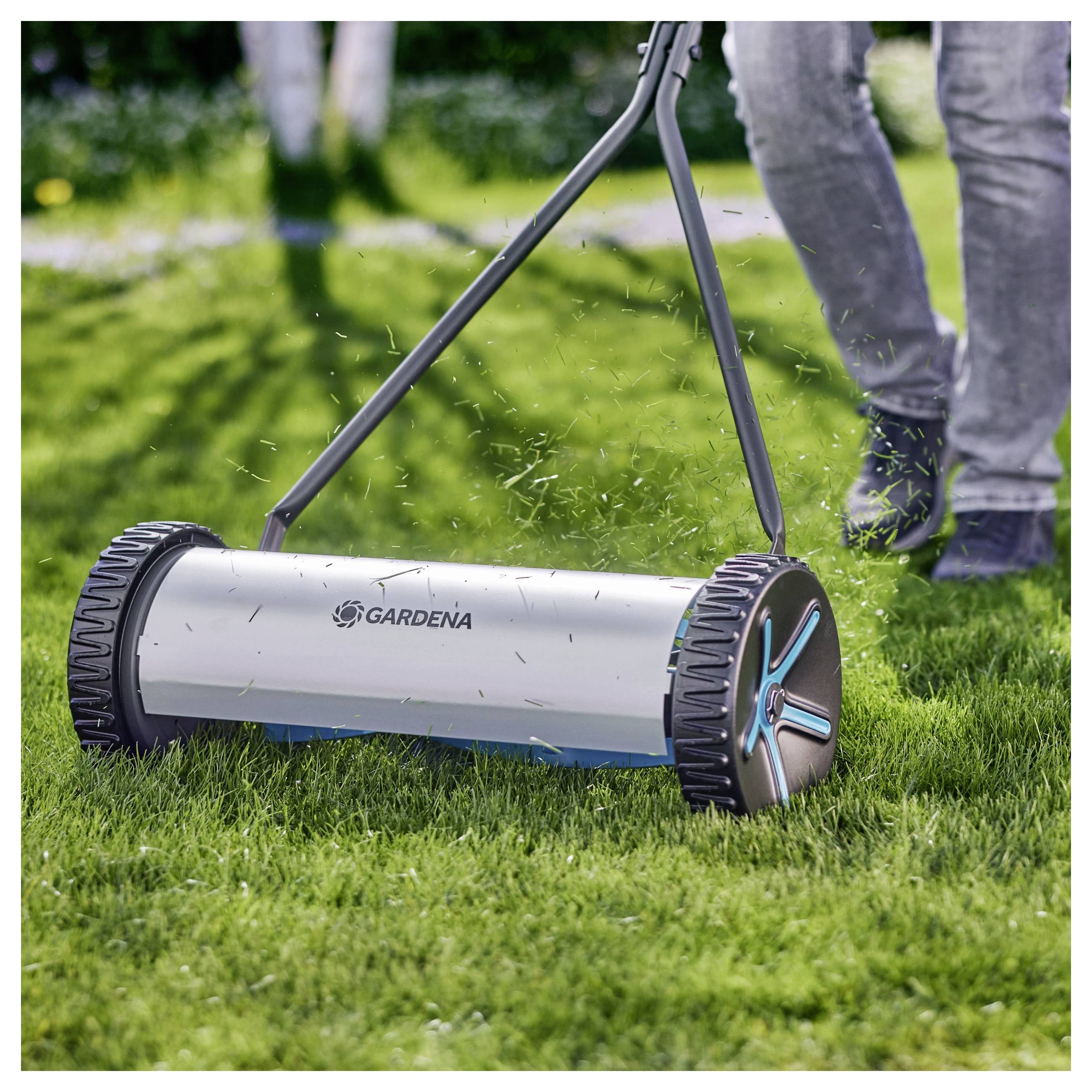 Handbetriebener Rasenmäher wird von einer Person über üppiges Gras geschoben, wobei der Fokus auf das moderne Design und die Funktionalität des Mähers liegt.