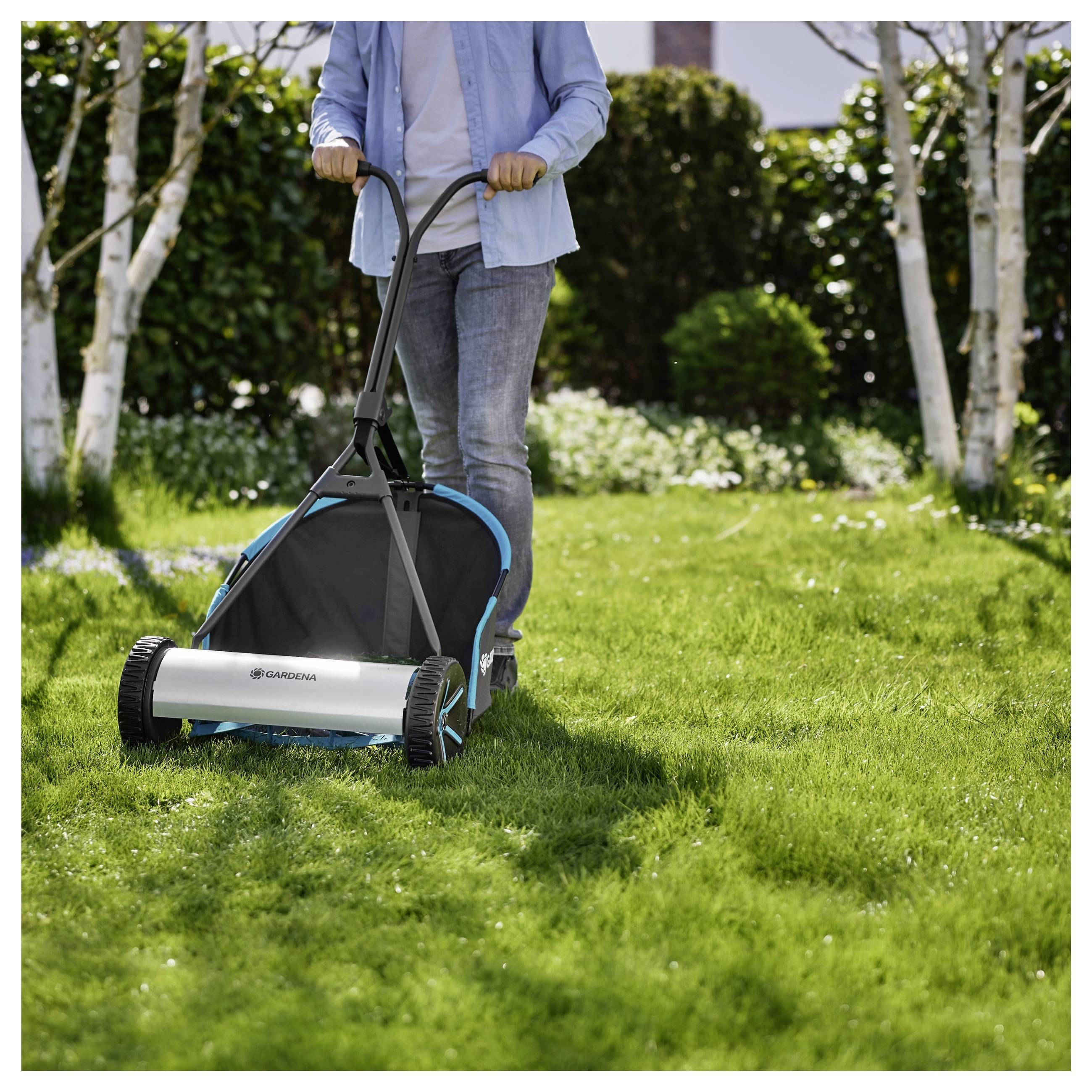 Eine Person benutzt einen manuellen Rasenmäher an einem sonnigen Tag. Der Mäher überquert eine grasbewachsene Fläche, mit Bäumen und Sträuchern im Hintergrund.