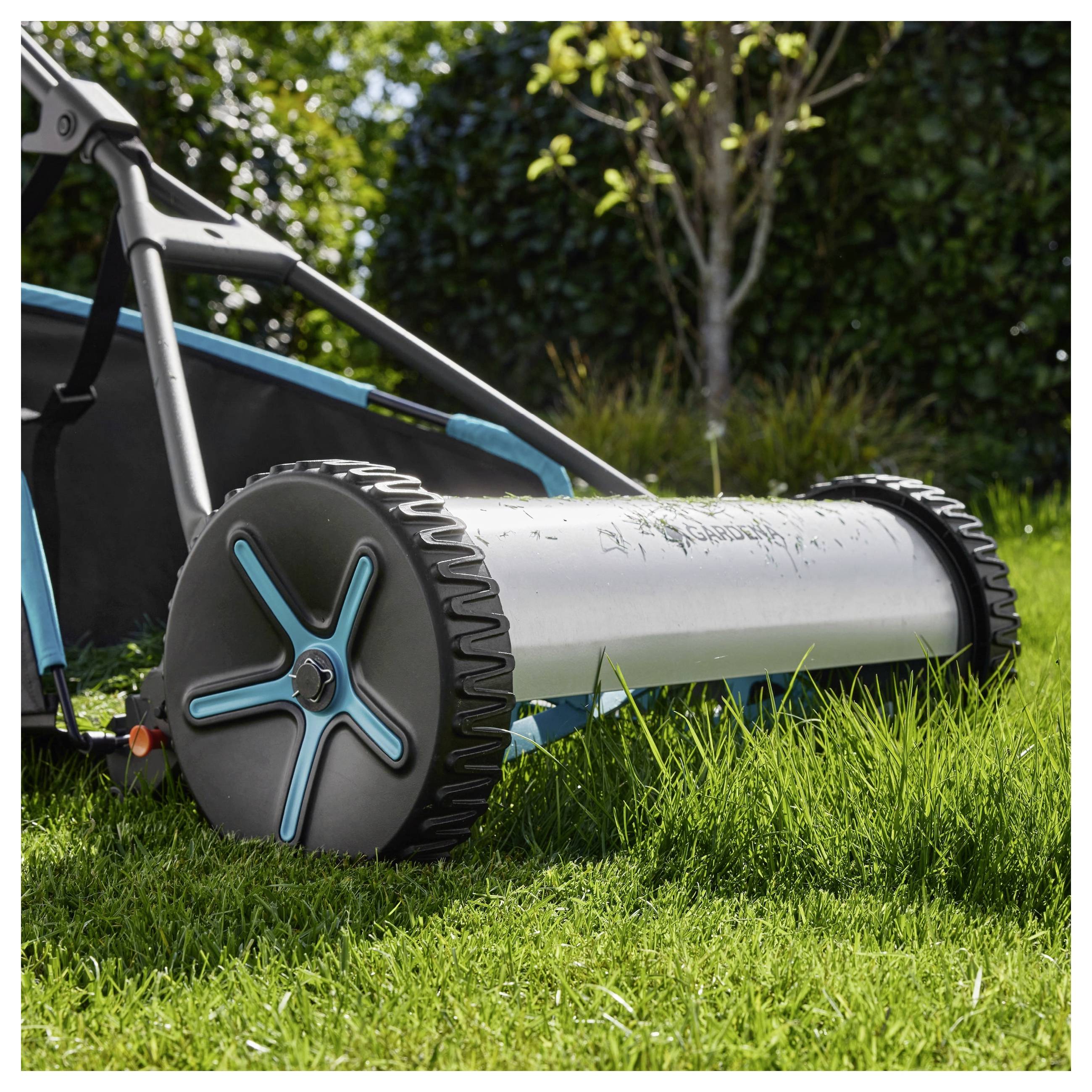 Ein manueller Rasenmäher mit blauen und silbernen Elementen schneidet Gras an einem sonnigen Tag, mit Bäumen und einer Hecke im Hintergrund.