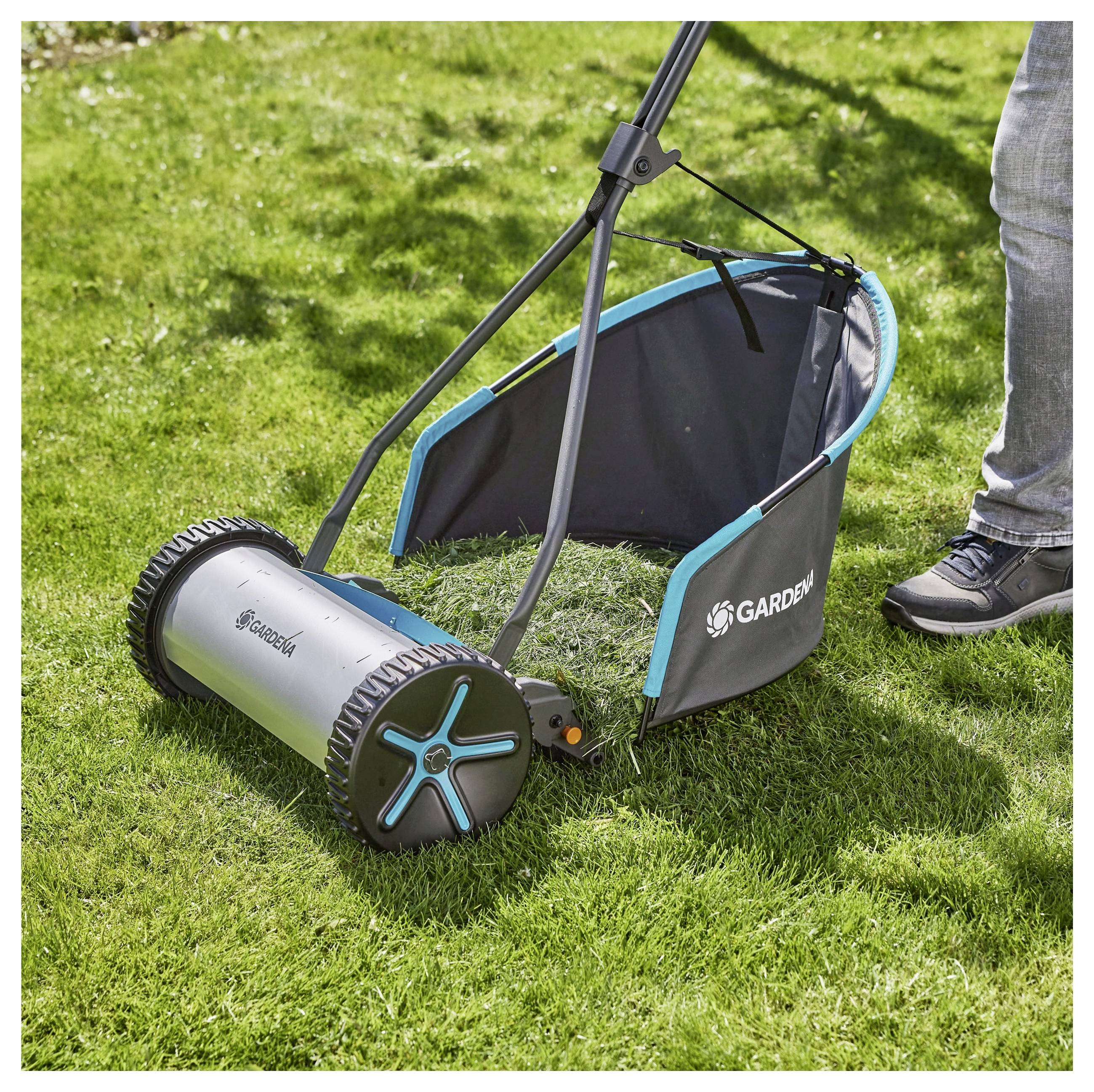 Ein manueller Rasenmäher mit Fangkorb; eine Person schiebt ihn auf einem grünen Rasen.